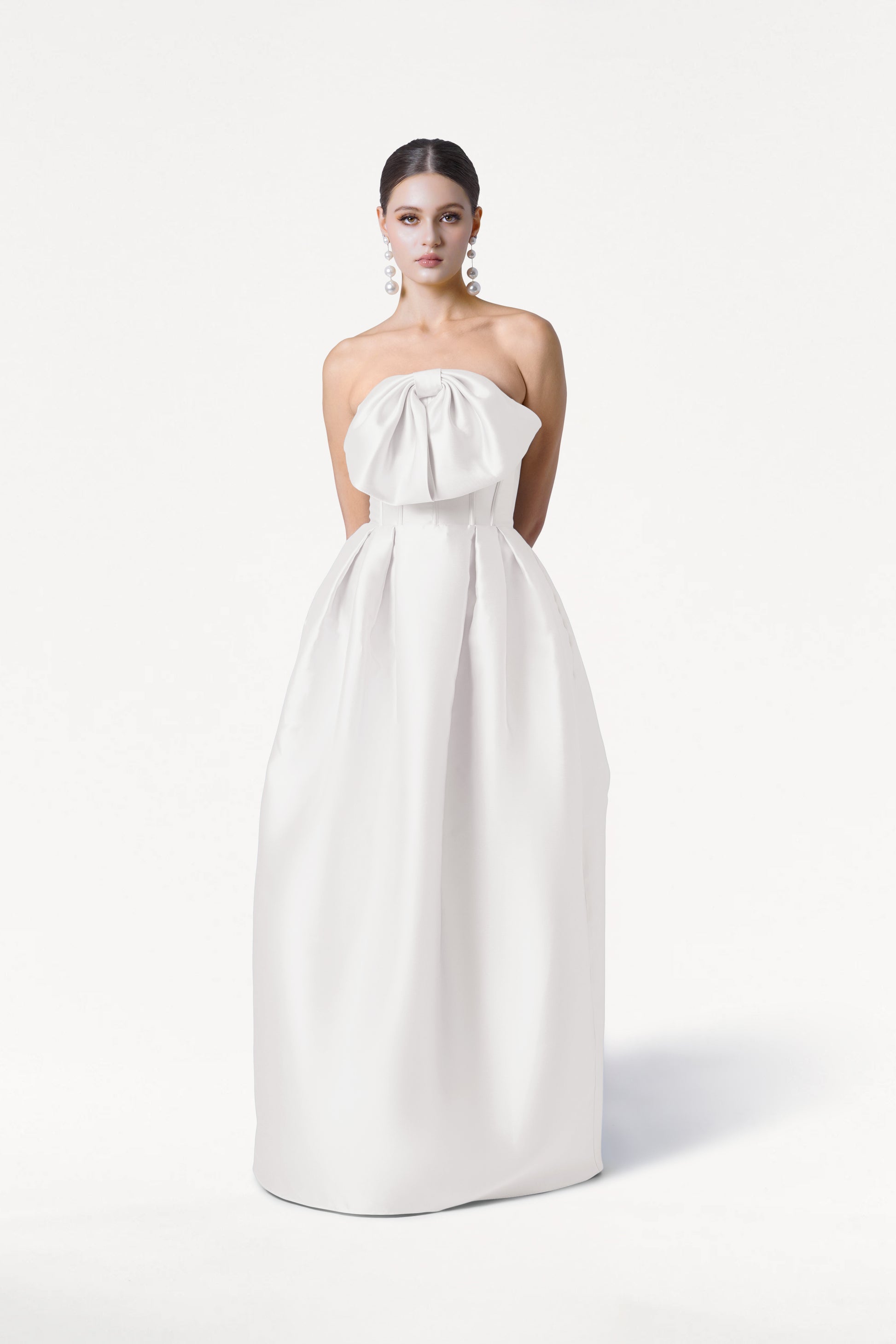 A woman stands against a white background wearing the TH&TH Ophelia Dress in Ivory Satin Twill, featuring a strapless neckline, large bow detail, and full floor-length skirt. She pairs it with pearl drop earrings and has her hair styled back.