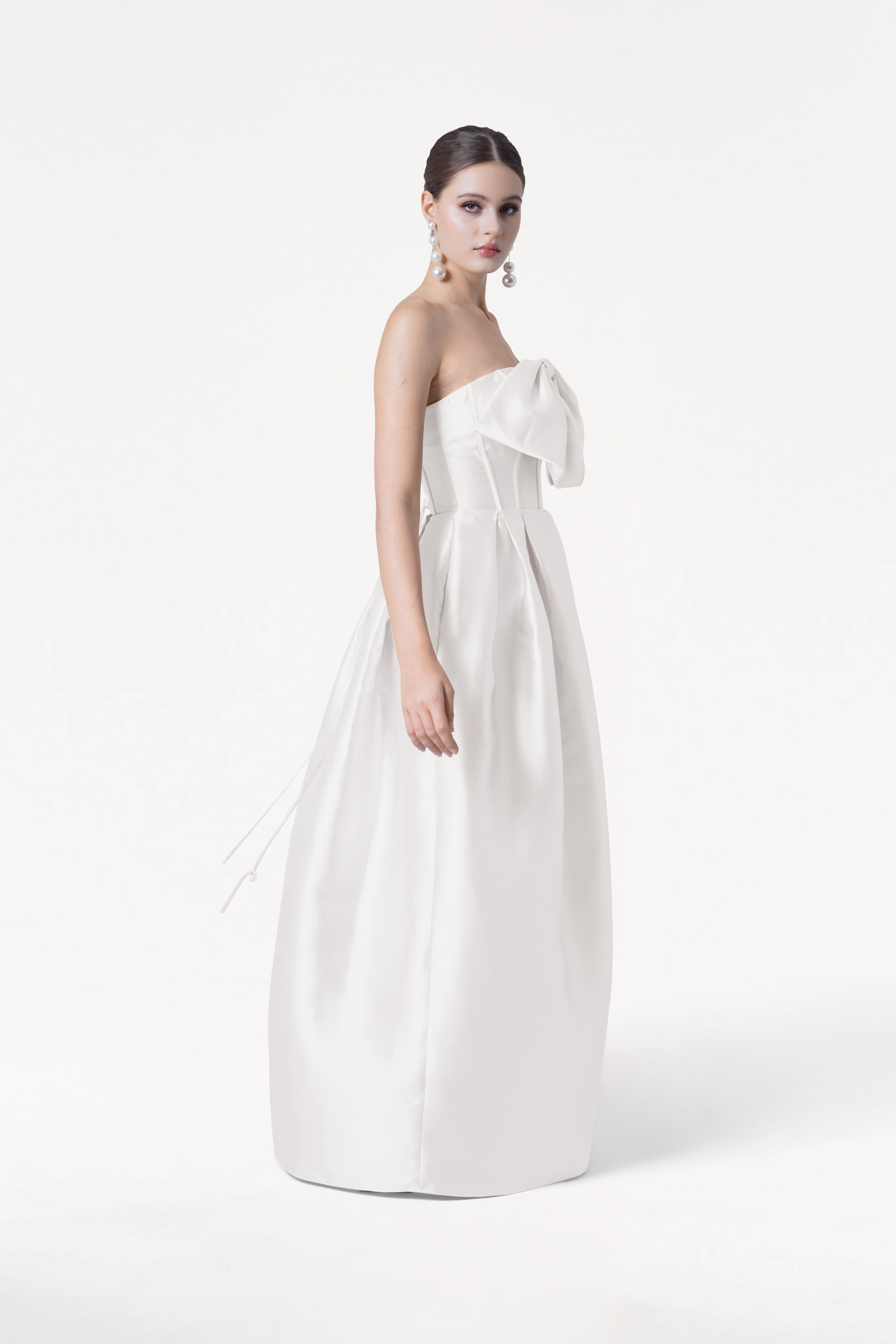 A woman stands against a white background wearing the TH&TH Ophelia Dress in Ivory Satin Twill, a strapless floor-length gown with a large bow on the bodice. Her hair is pulled back, and she wears statement earrings.
