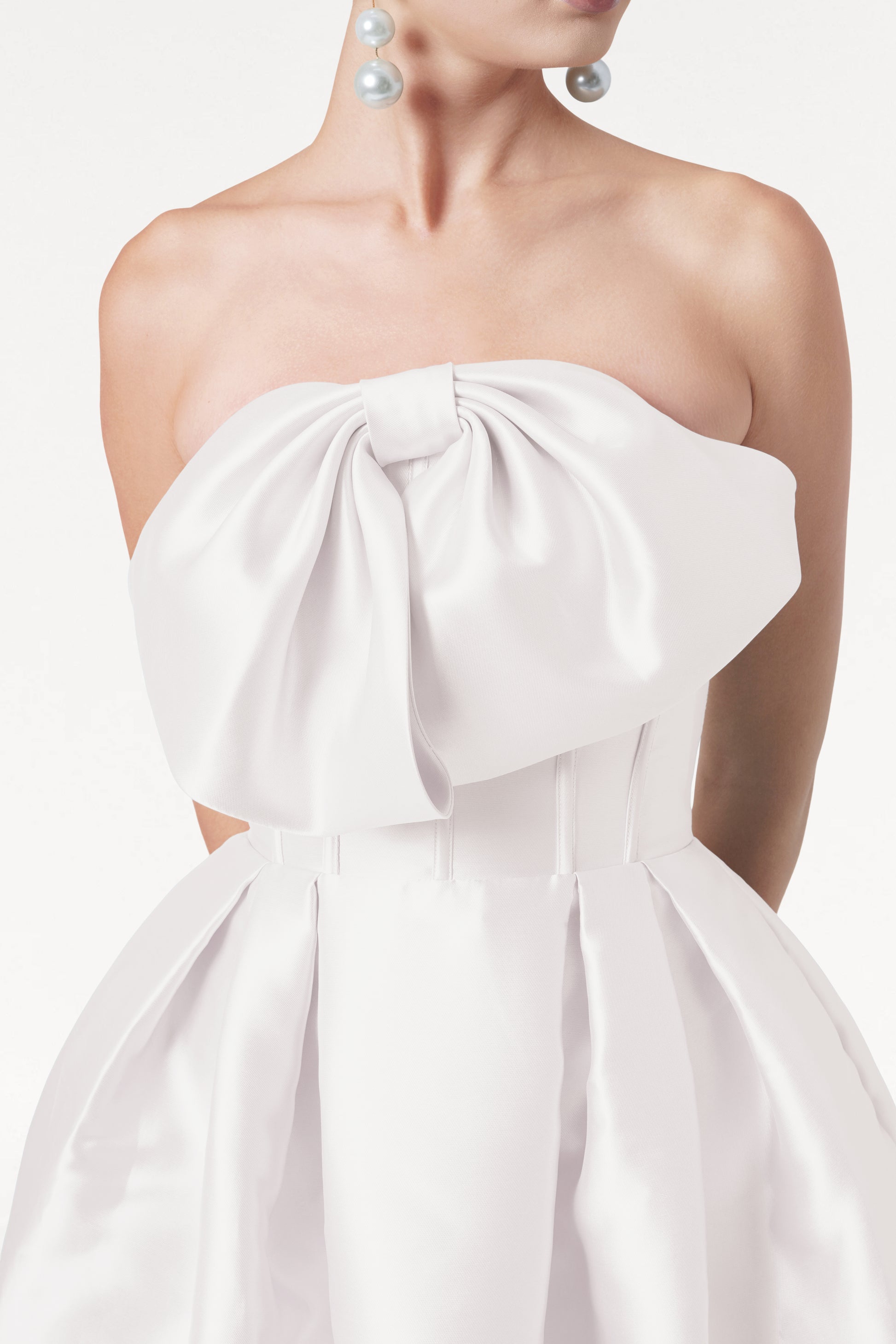 A woman wears the TH&TH Ophelia Dress in Ivory Satin Twill, featuring a strapless fitted corset bodice and a large bow. She pairs it with statement pearl drop earrings. The image is cropped from shoulders to above the knees.