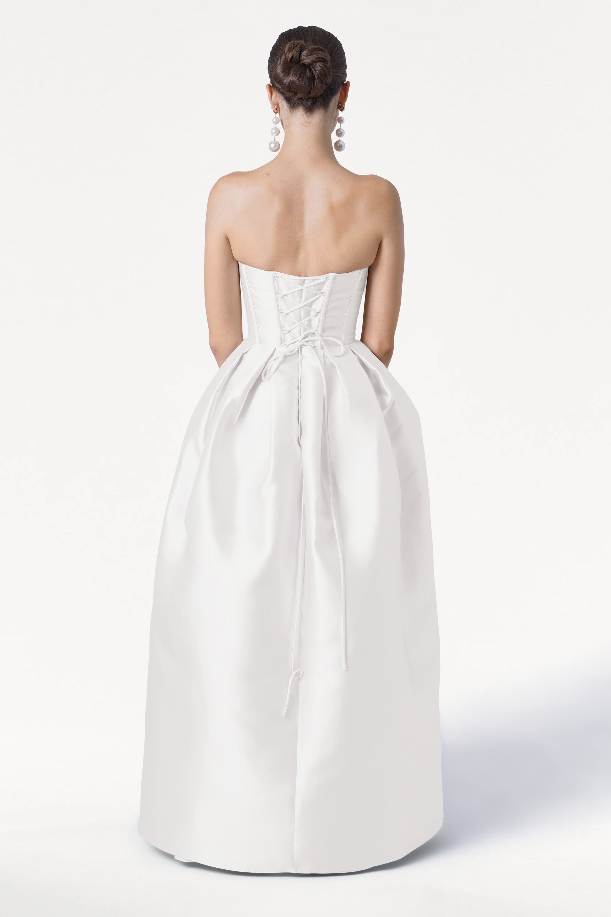 A woman with dark hair in a bun faces away from the camera against a white background, wearing the TH&TH Ophelia Dress in Ivory Satin Twill—a strapless gown with a corset bodice and full skirt—and dangling silver earrings.