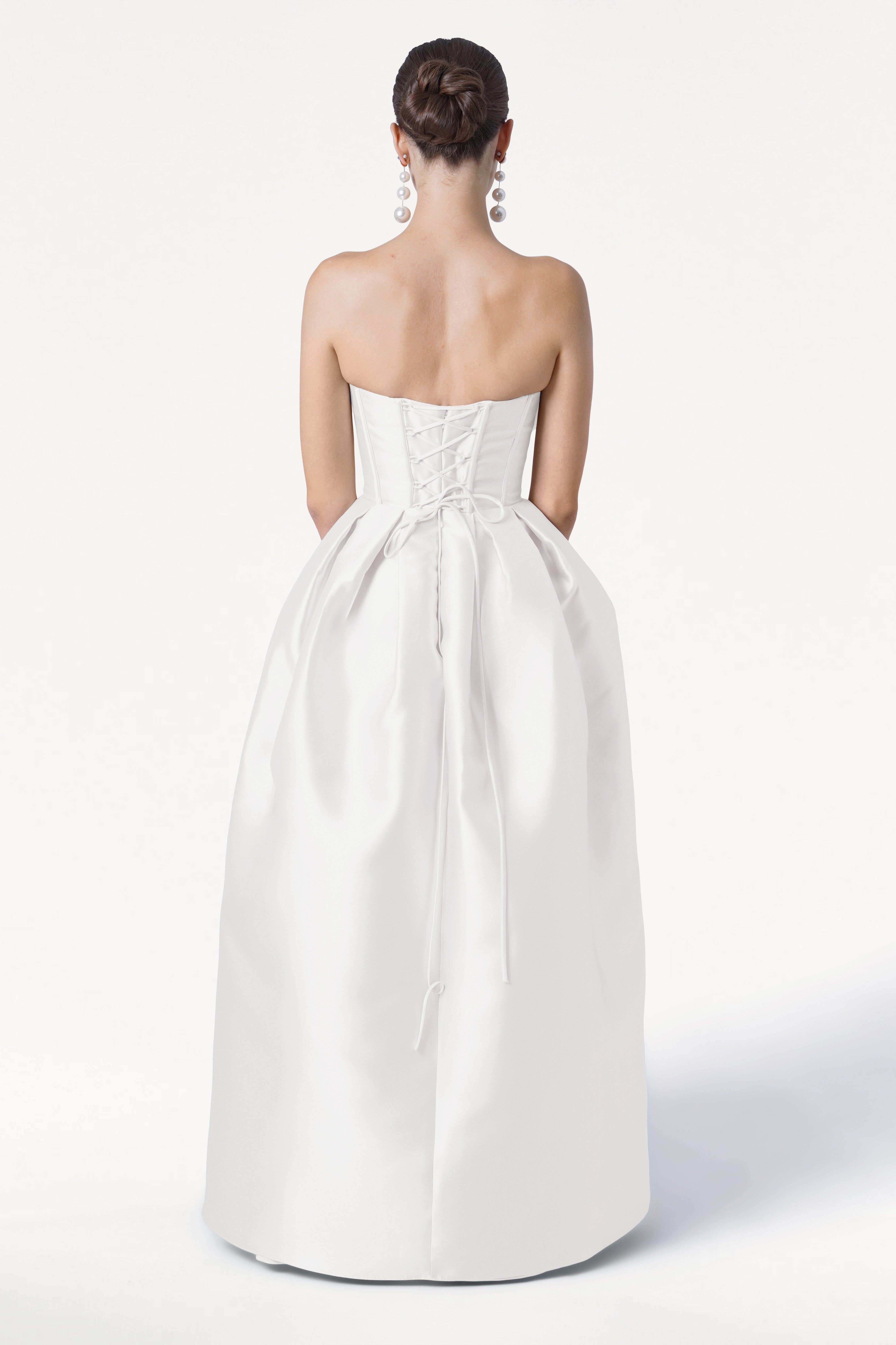 A woman with dark hair in a bun faces away from the camera against a white background, wearing the TH&TH Ophelia Dress in Ivory Satin Twill—a strapless gown with a corset bodice and full skirt—and dangling silver earrings.