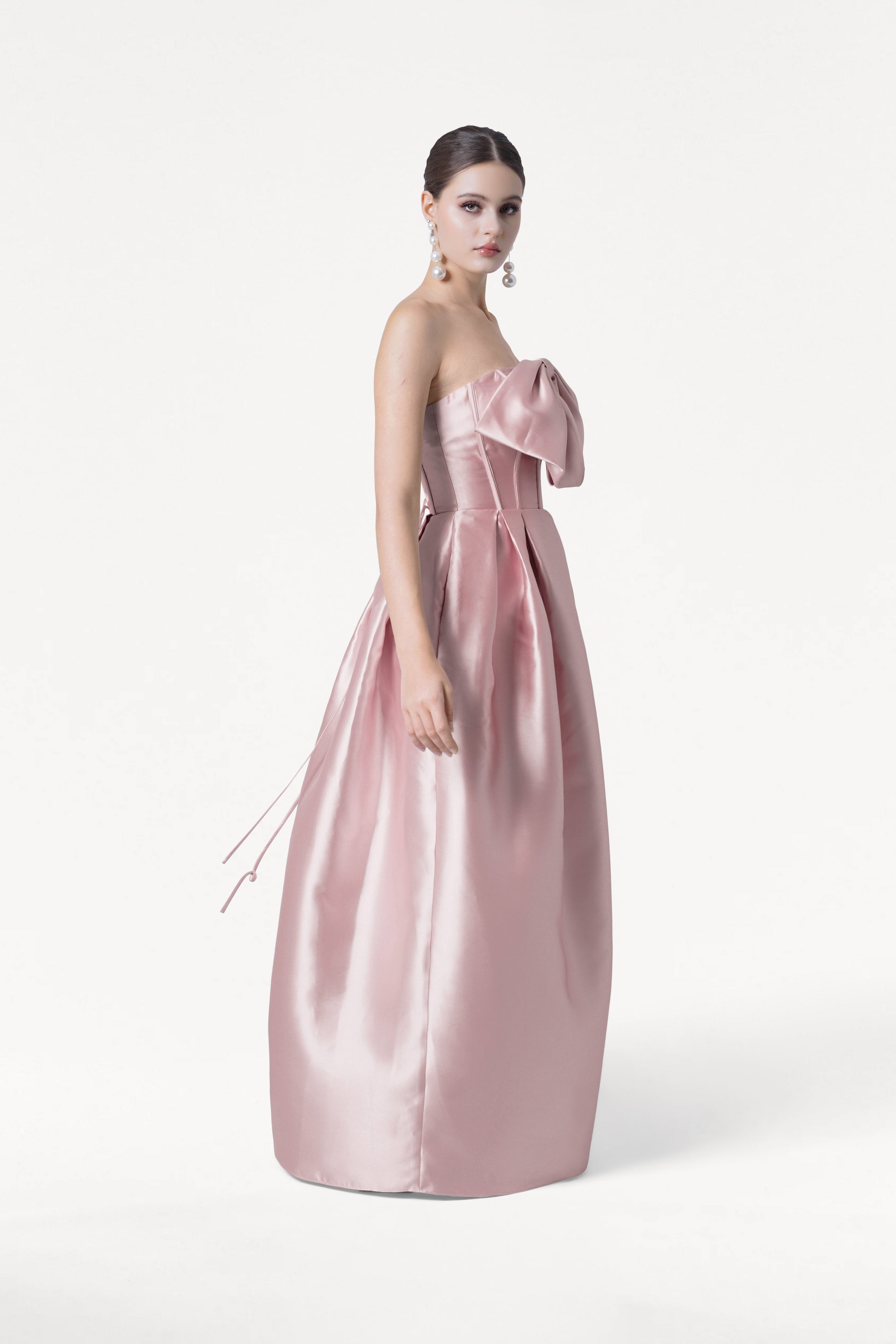 A woman poses against a white background in the TH&TH Ophelia Bridesmaid Dress, a strapless, floor-length smoked blush satin twill gown featuring a large bow on the corset bodice. She wears dangly earrings with her dark hair neatly styled back.