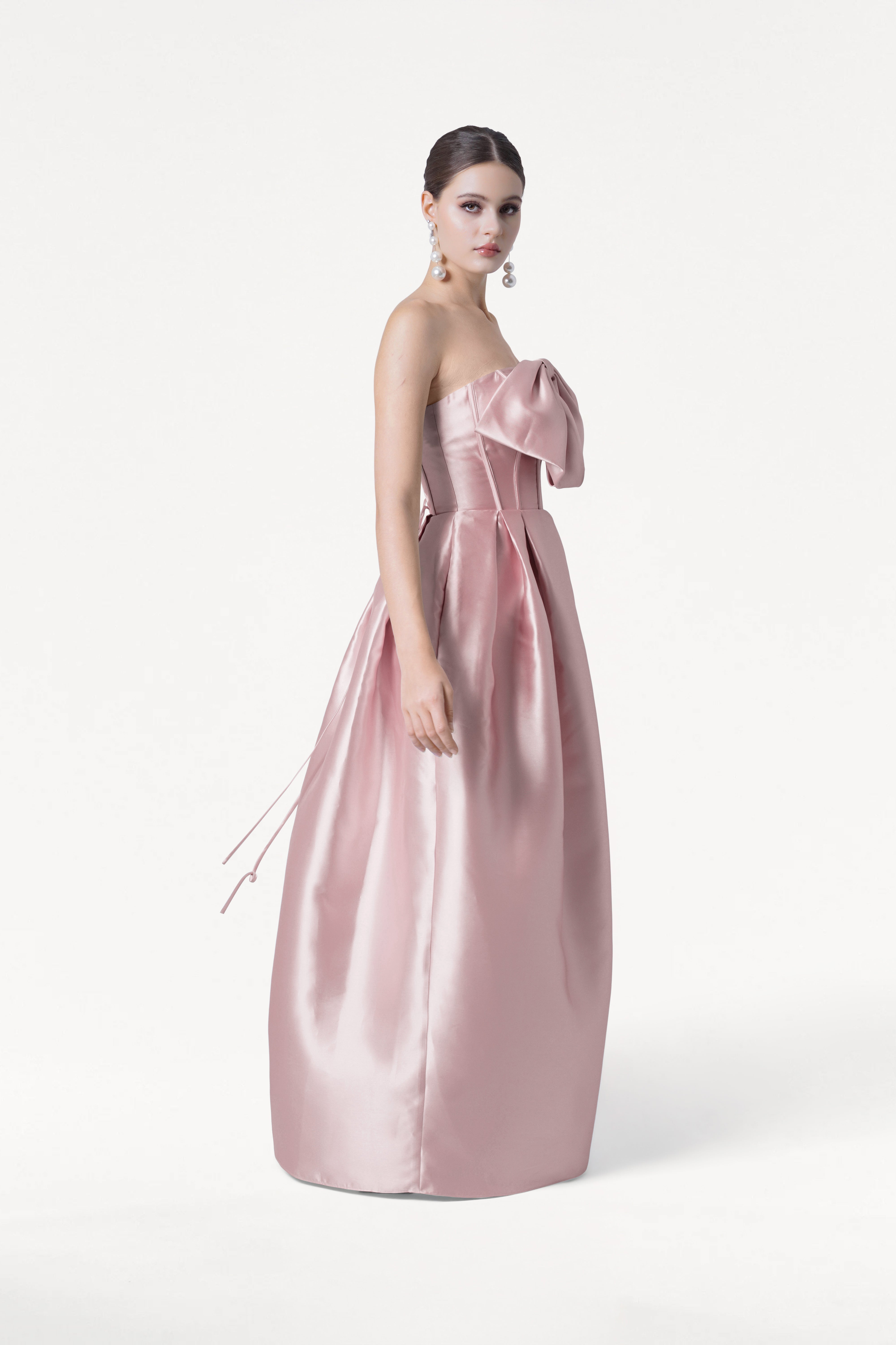 A woman poses against a white background in the TH&TH Ophelia Bridesmaid Dress, a strapless, floor-length smoked blush satin twill gown featuring a large bow on the corset bodice. She wears dangly earrings with her dark hair neatly styled back.