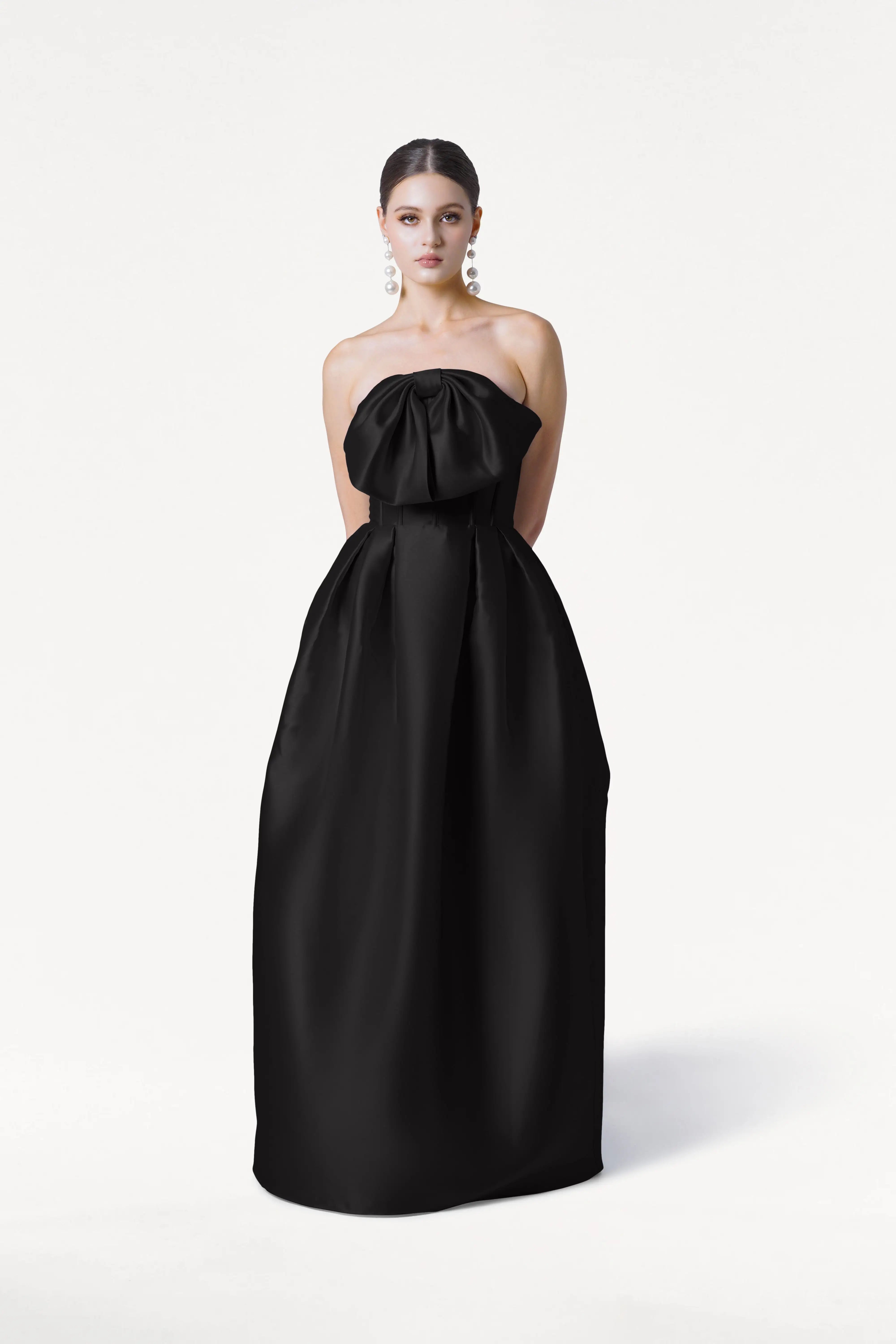A woman stands against a white background wearing the TH&TH Ophelia Bridesmaid Dress in Black Satin Twill, a strapless, floor-length gown featuring a large bow at the corset bodice. Her hair is pulled back and she wears dangling earrings.