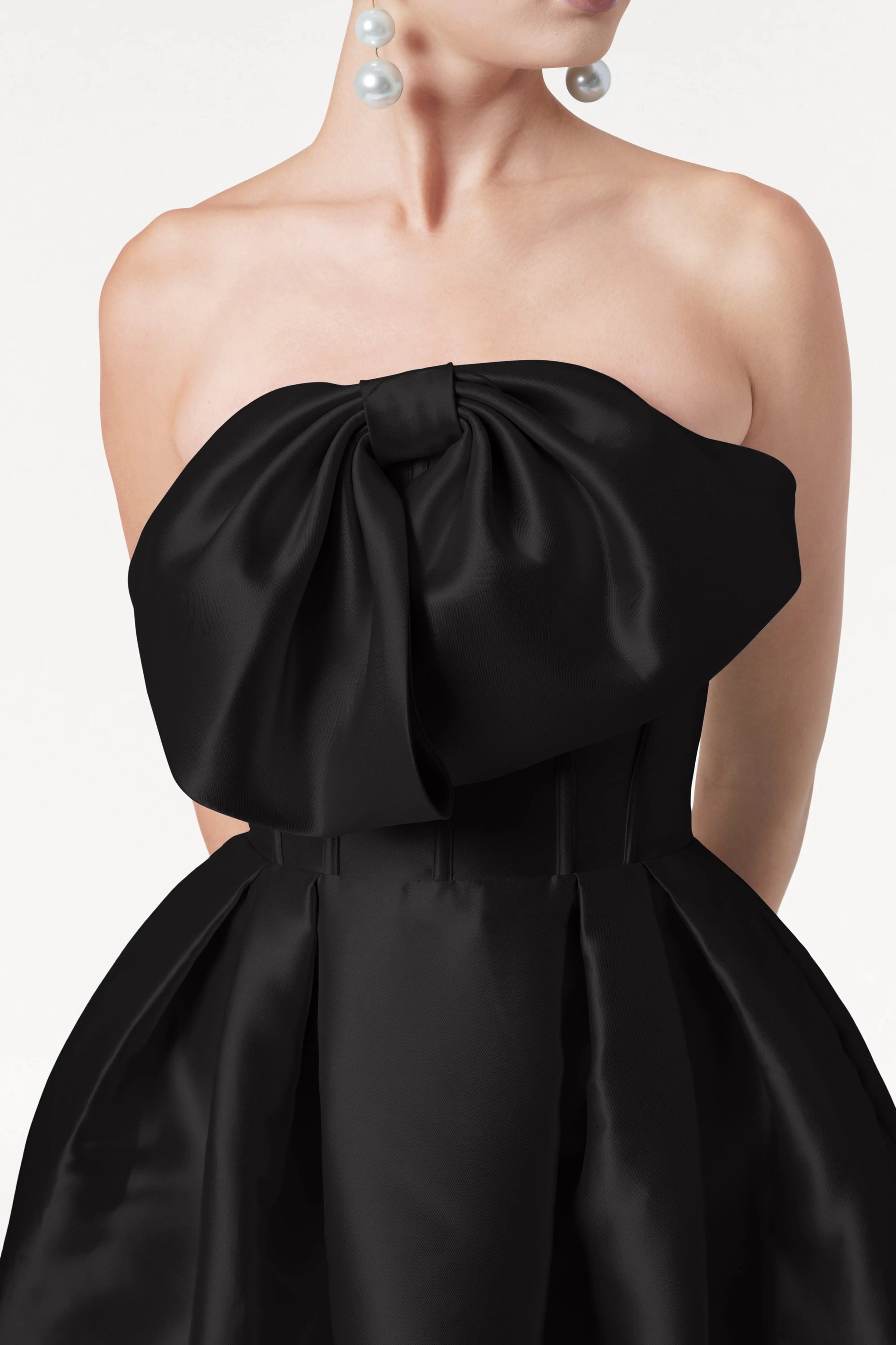 A woman wears the TH&TH Ophelia Bridesmaid Dress in Black Satin Twill with a corset bodice and large bow, paired with oversized pearl drop earrings. Her head is partially out of frame against a plain white background.