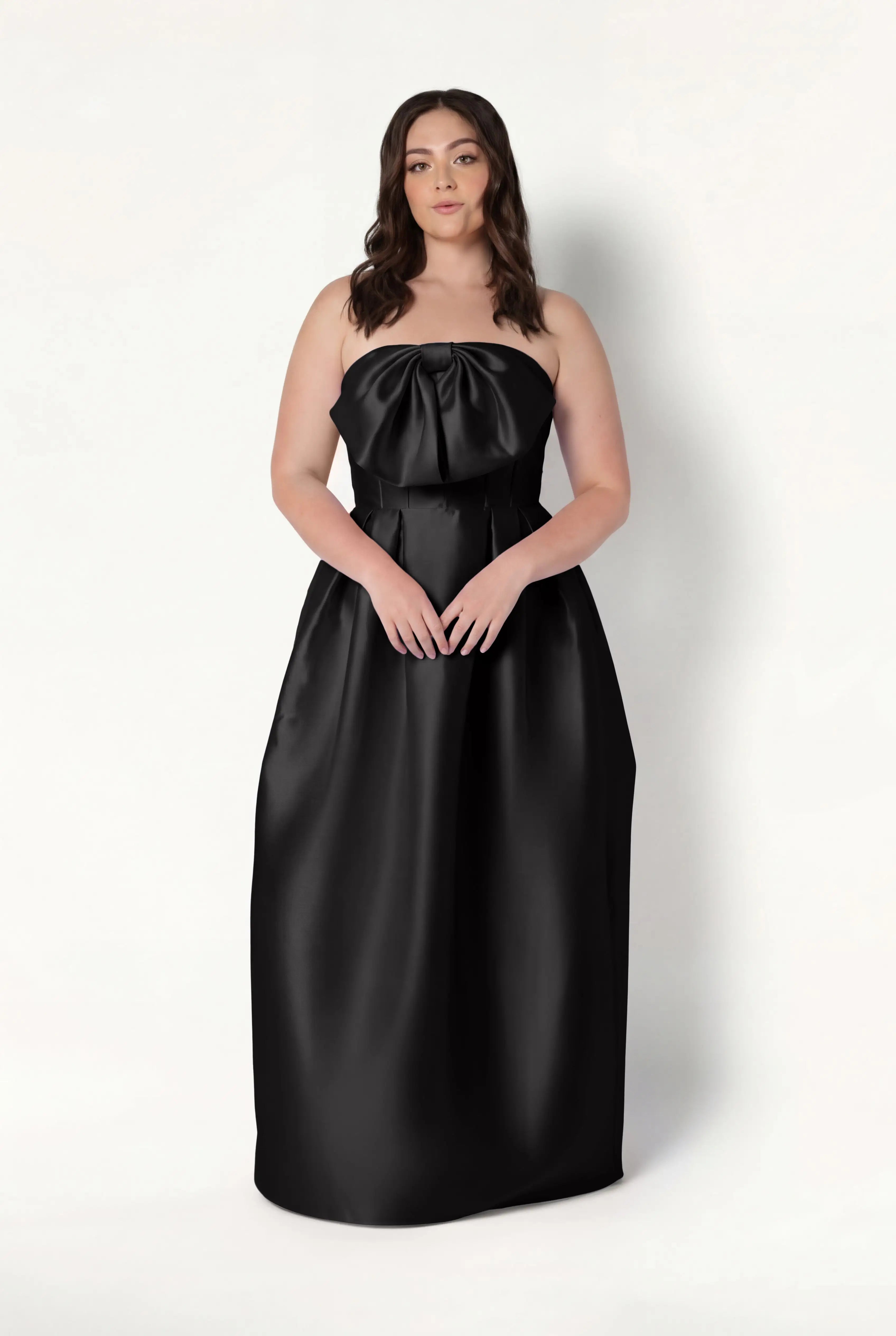A woman with long brown hair models the TH&TH Ophelia Bridesmaid Dress in Black Satin Twill—a strapless, floor-length gown with a corset bodice and large bow at the neckline—against a white background.