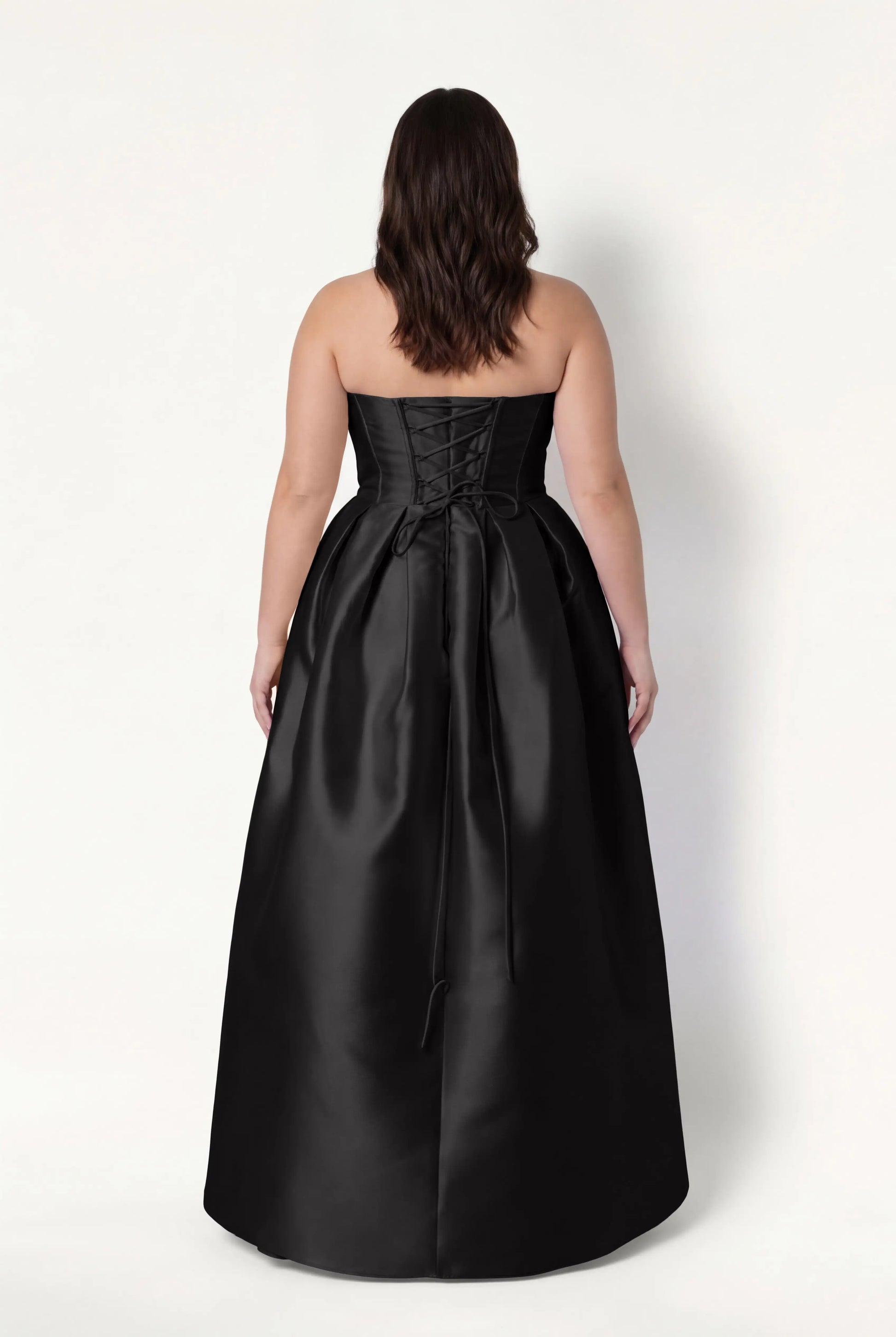 A woman with wavy brown hair models the TH&TH Ophelia Bridesmaid Dress in Black Satin Twill, a strapless, floor-length gown with a corset lace-up back, standing against a plain white background and facing away from the camera.