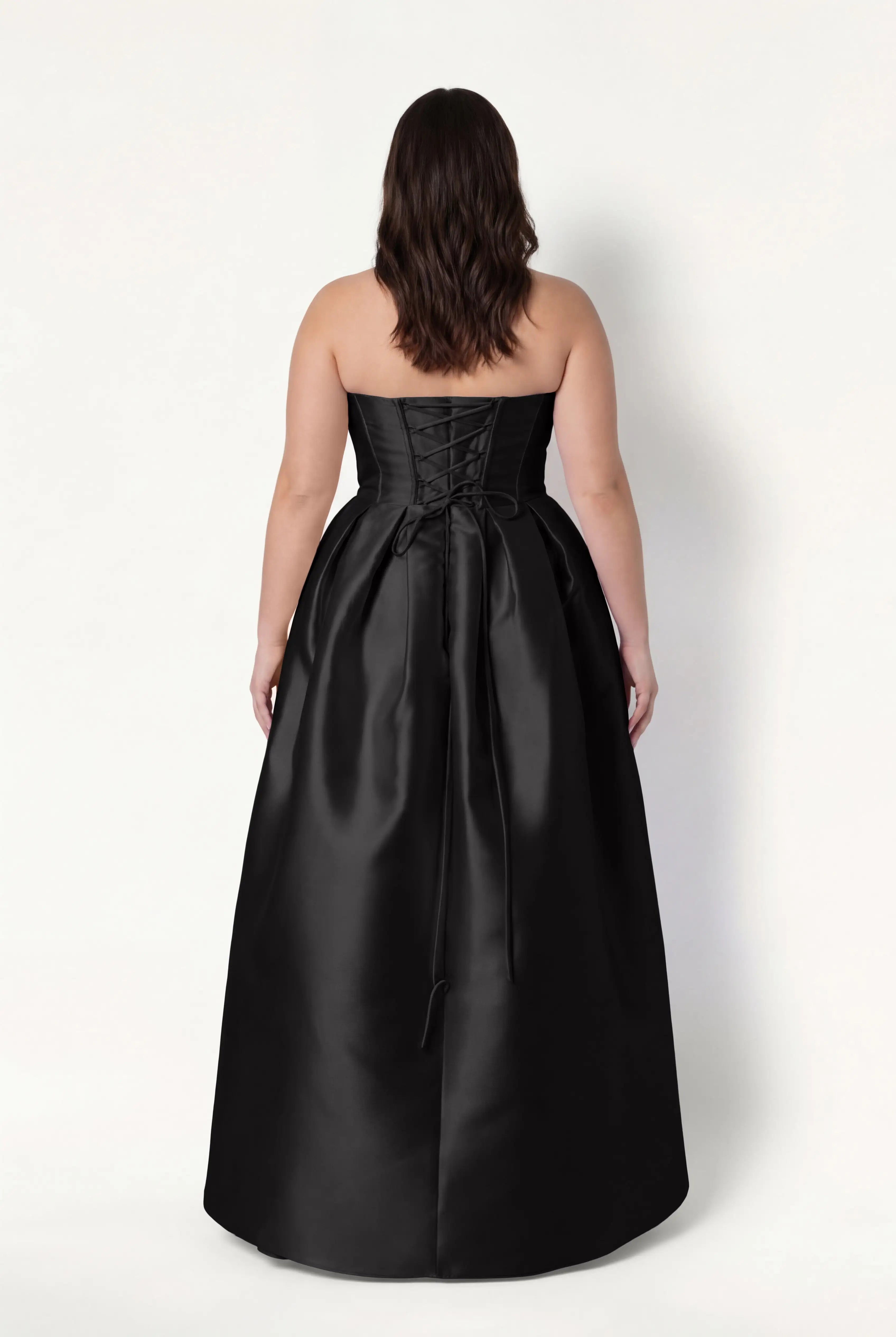 A woman with wavy brown hair models the TH&TH Ophelia Bridesmaid Dress in Black Satin Twill, a strapless, floor-length gown with a corset lace-up back, standing against a plain white background and facing away from the camera.