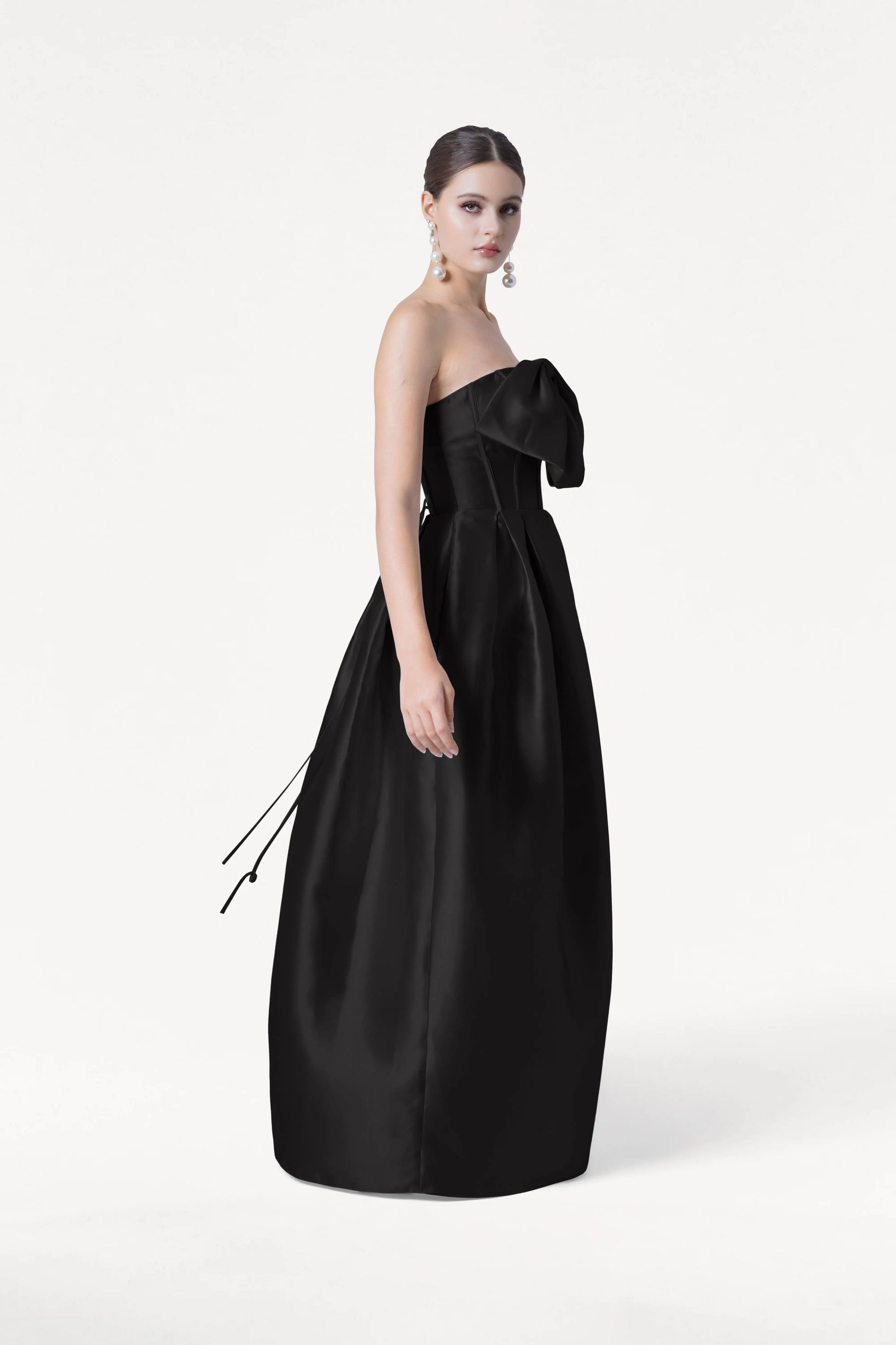 A woman stands against a white background, wearing the TH&TH Ophelia Bridesmaid Dress in black satin twill, a strapless floor-length gown with a large bow on the corset bodice. Her hair is pulled back and she wears dangling earrings.