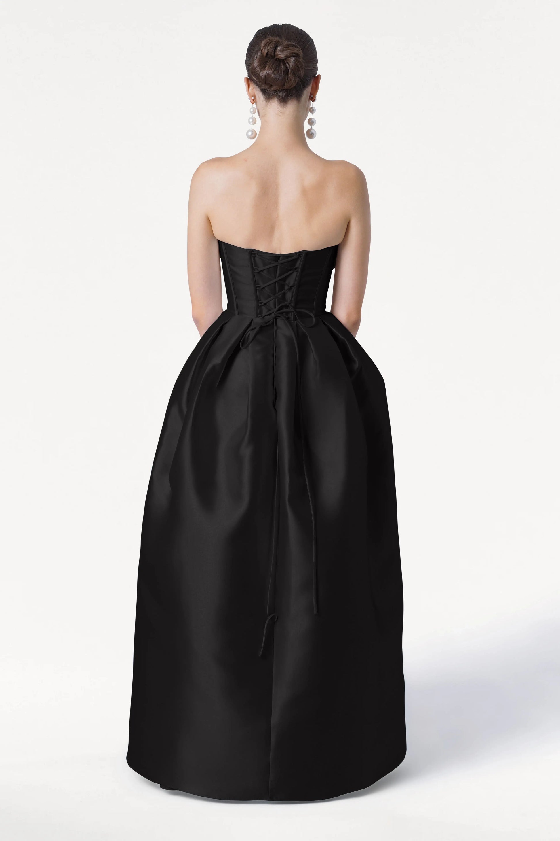 A woman with dark hair in a neat bun wears TH&TH’s Ophelia Bridesmaid Dress in Black Satin Twill, featuring a strapless lace-up corset bodice and bow. She faces away, showcasing its elegant floor-length design with large dangling earrings.
