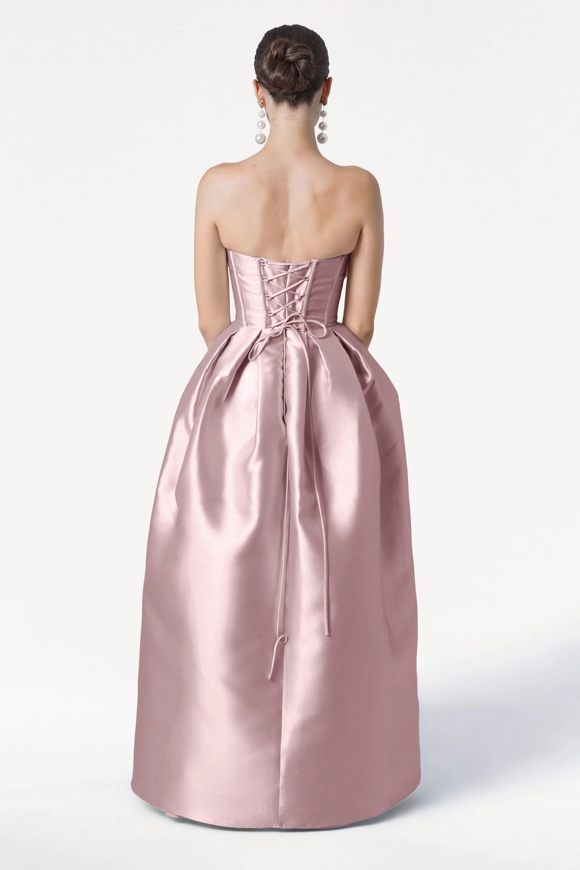 A woman with a neat bun and long, dangling earrings stands with her back to the camera, wearing the TH&TH Ophelia Bridesmaid Dress in blush pink satin twill, featuring a strapless design and lace-up corset bodice.