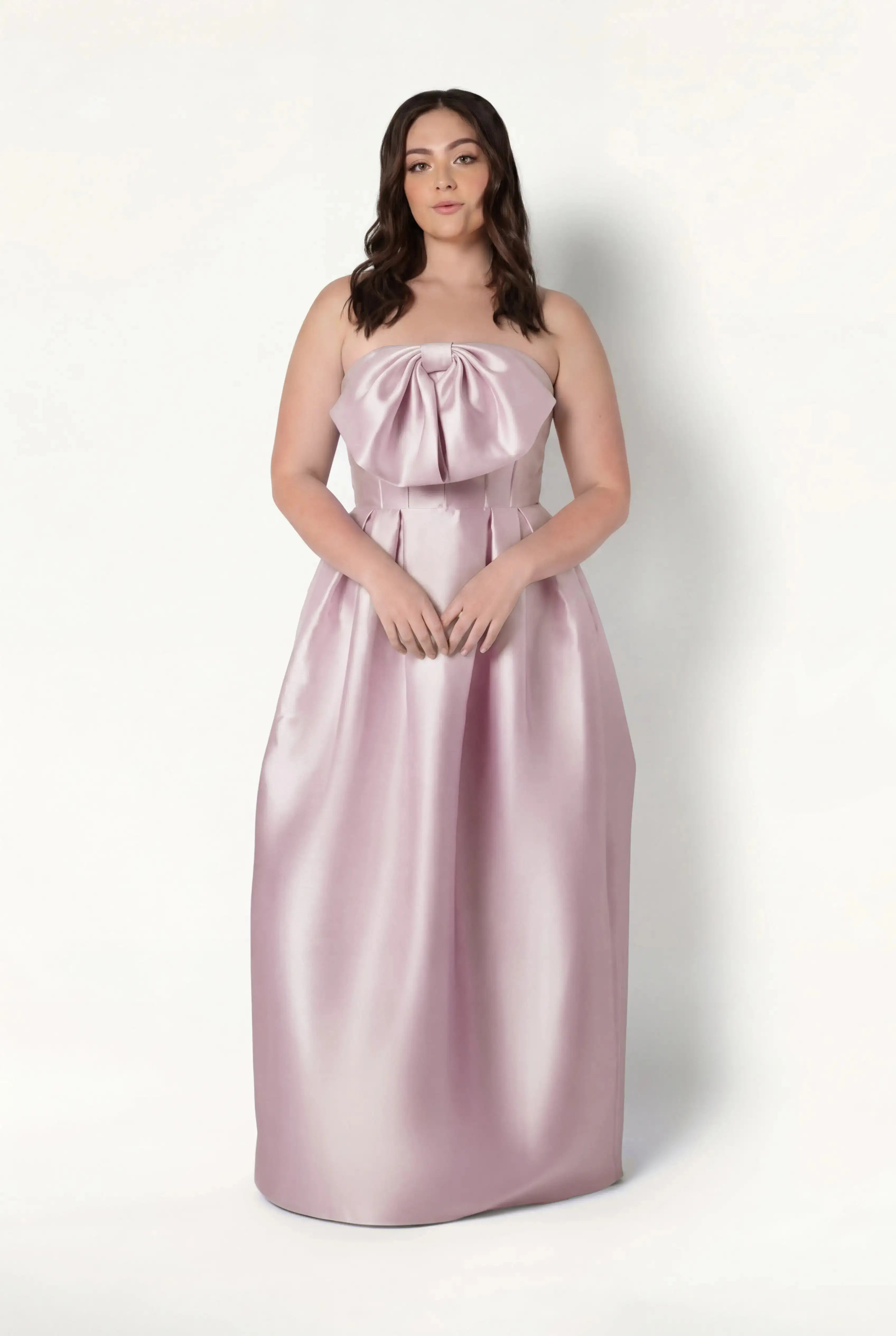A woman with wavy brown hair wears the TH&TH Ophelia Bridesmaid Dress in Blush Pink Satin Twill, featuring a strapless design and large bow on the bodice, while posing confidently against a plain white background.