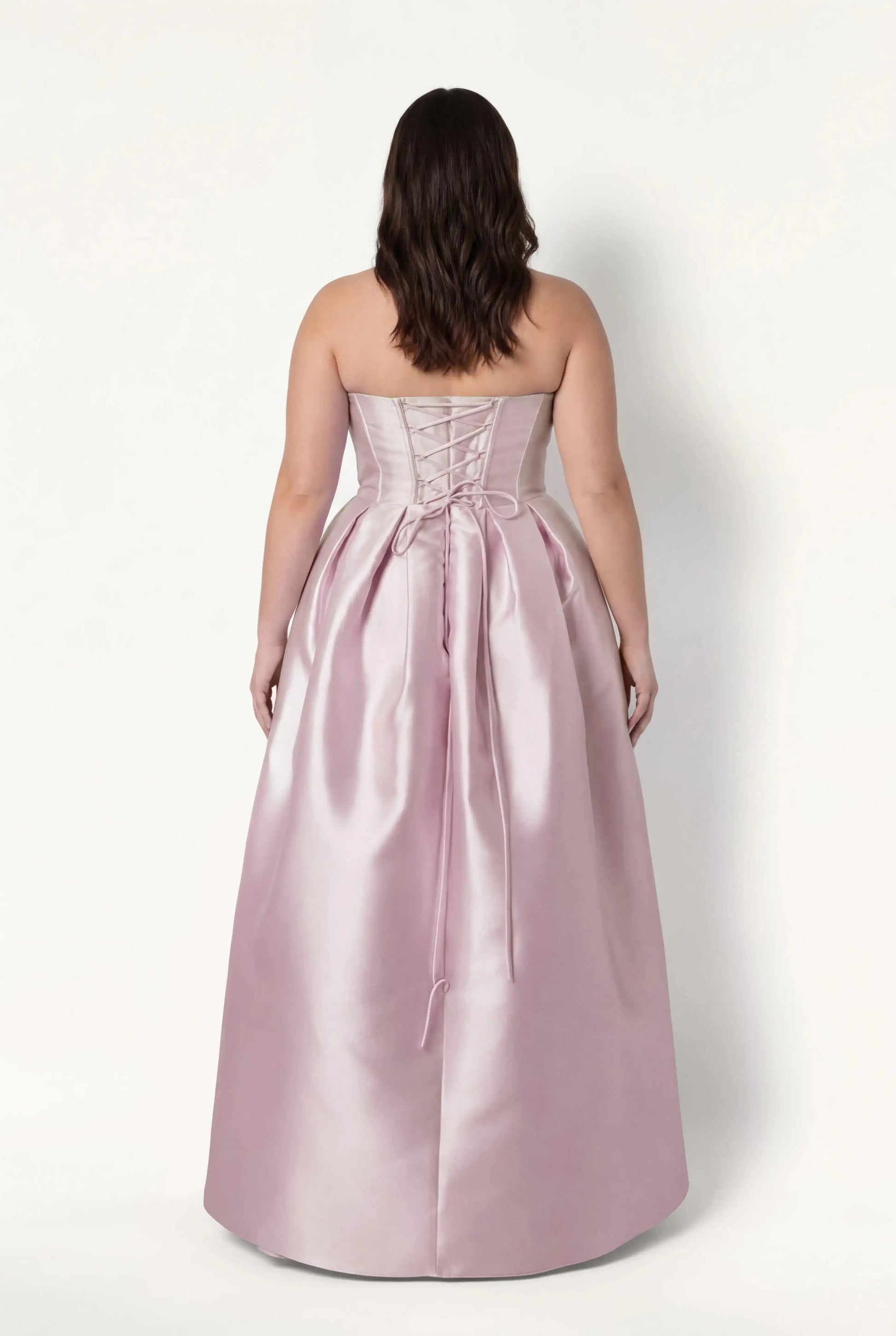 A woman with wavy brown hair models the TH&TH Ophelia Bridesmaid Dress in Blush Pink Satin Twill, featuring a strapless lace-up corset bodice and gathered floor-length skirt, standing against a plain white background, facing away.