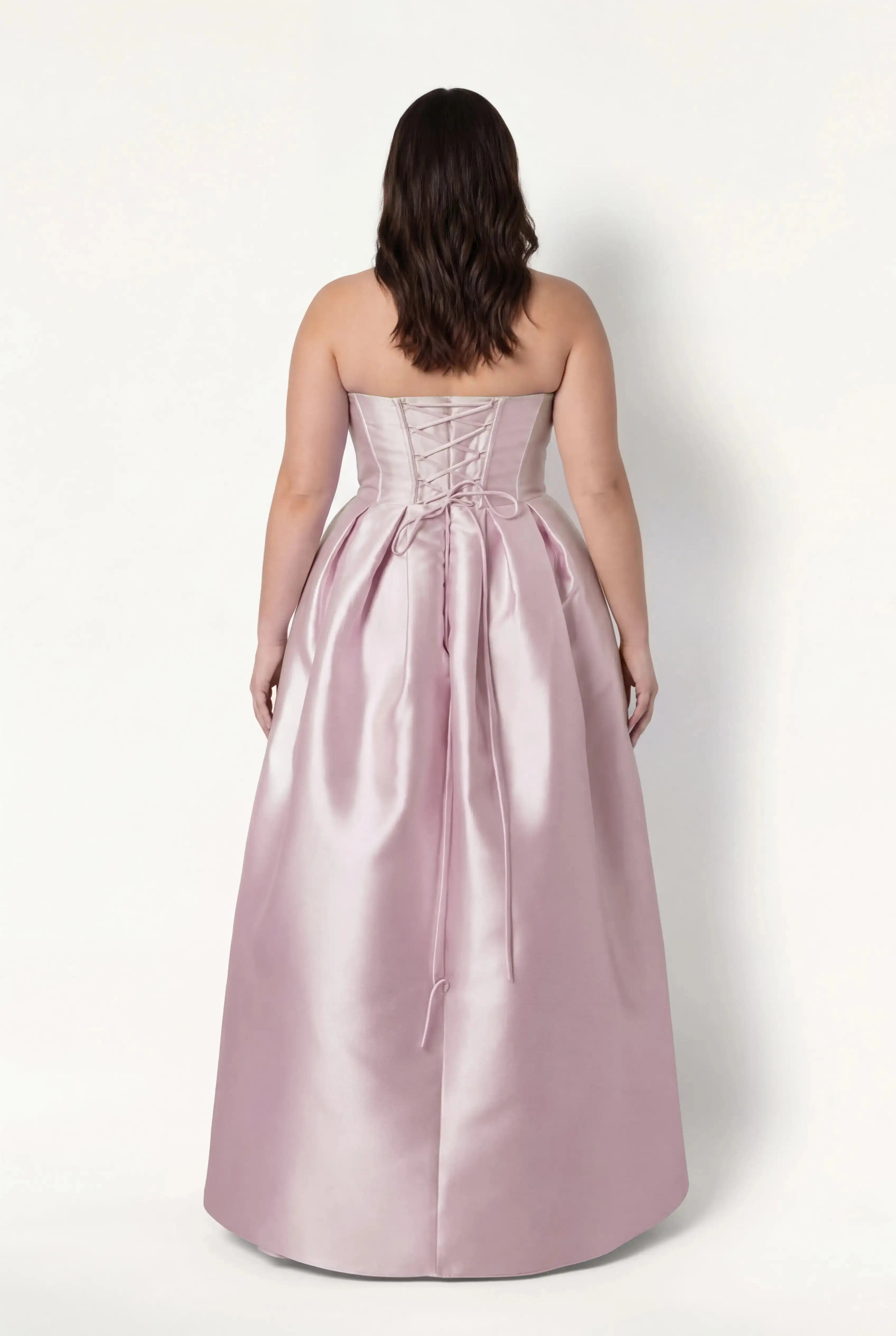 A woman with wavy brown hair models the TH&TH Ophelia Bridesmaid Dress in Blush Pink Satin Twill, featuring a strapless lace-up corset bodice and gathered floor-length skirt, standing against a plain white background, facing away.