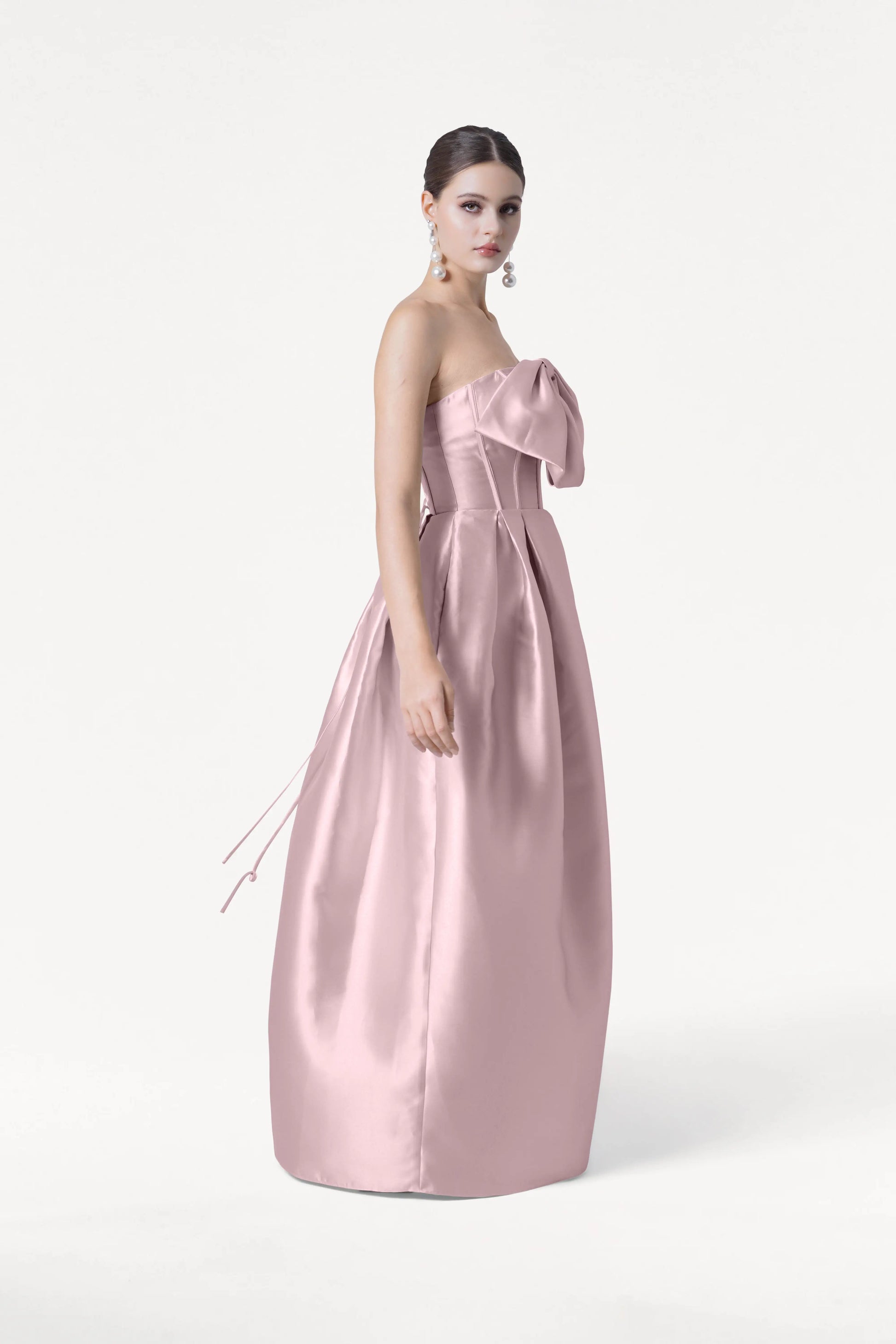 A woman models the Ophelia Bridesmaid Dress by TH&TH, a strapless, floor-length blush pink satin twill gown with a large bow on the corset bodice. She wears her hair up with dangling earrings and poses against a plain white background.