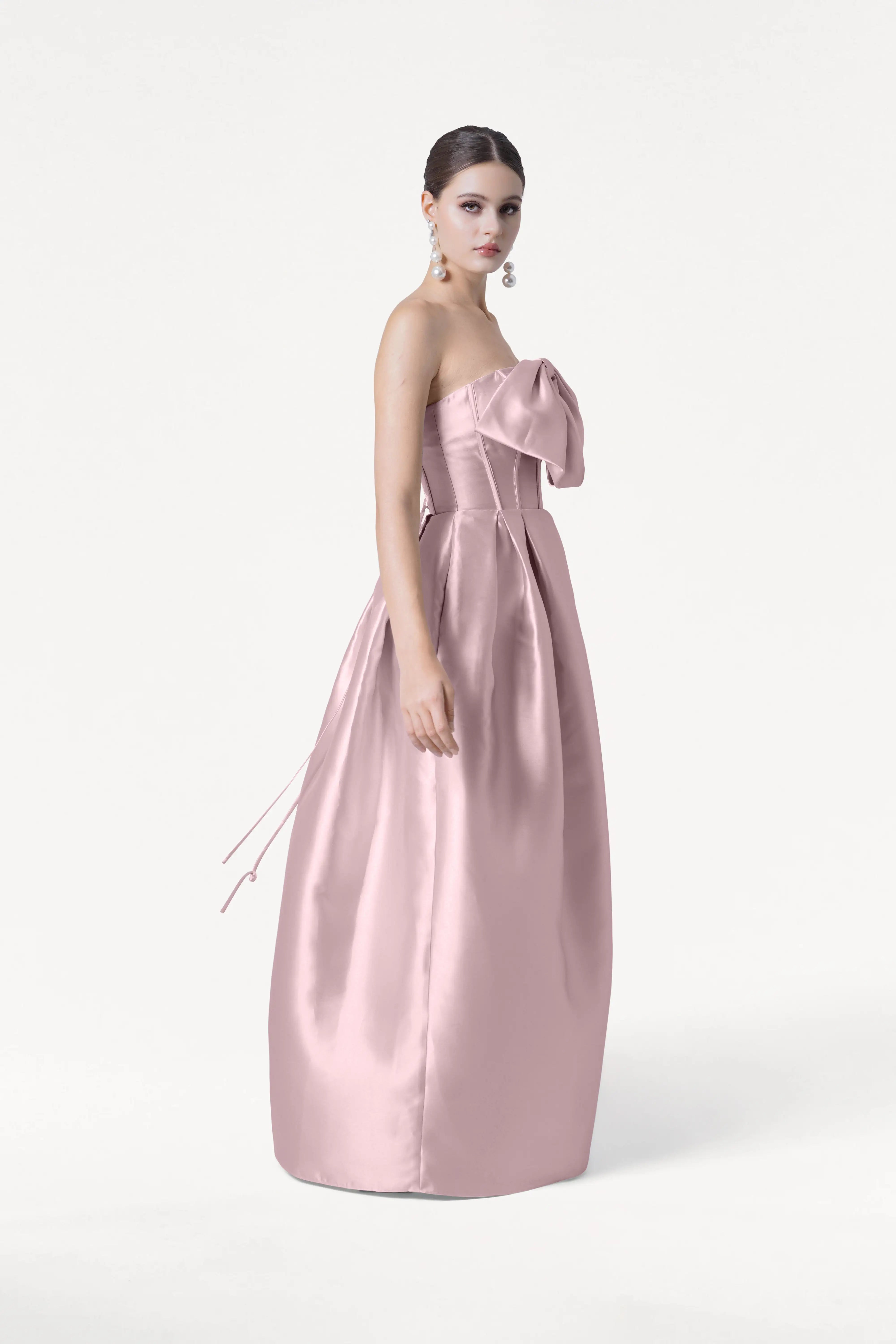 A woman models the Ophelia Bridesmaid Dress by TH&TH, a strapless, floor-length blush pink satin twill gown with a large bow on the corset bodice. She wears her hair up with dangling earrings and poses against a plain white background.