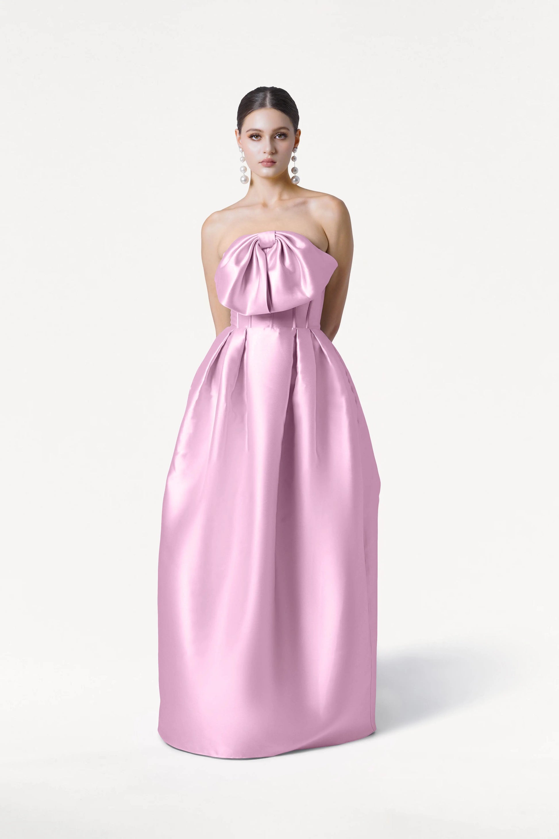 A woman models the Ophelia Bridesmaid Dress in Bubblegum Pink Satin Twill by TH&TH—a strapless gown with a corset bodice and large bow—against a white background, accessorized with statement earrings and hands behind her back.