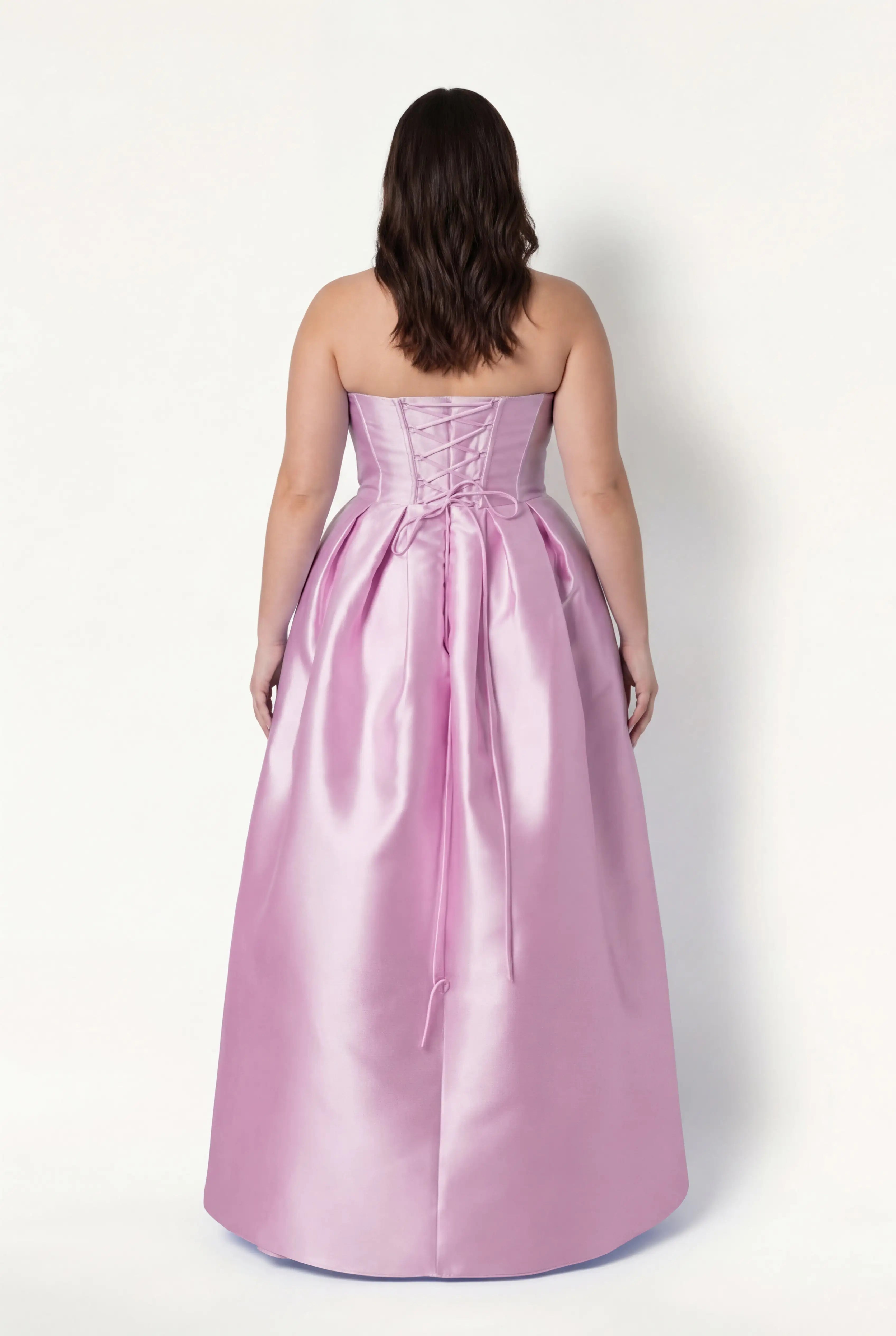A woman with wavy, shoulder-length brown hair models the TH&TH Ophelia Bridesmaid Dress in Bubblegum Pink Satin Twill, showing off its strapless, floor-length design and lace-up corset bodice as she stands with her back to the camera.