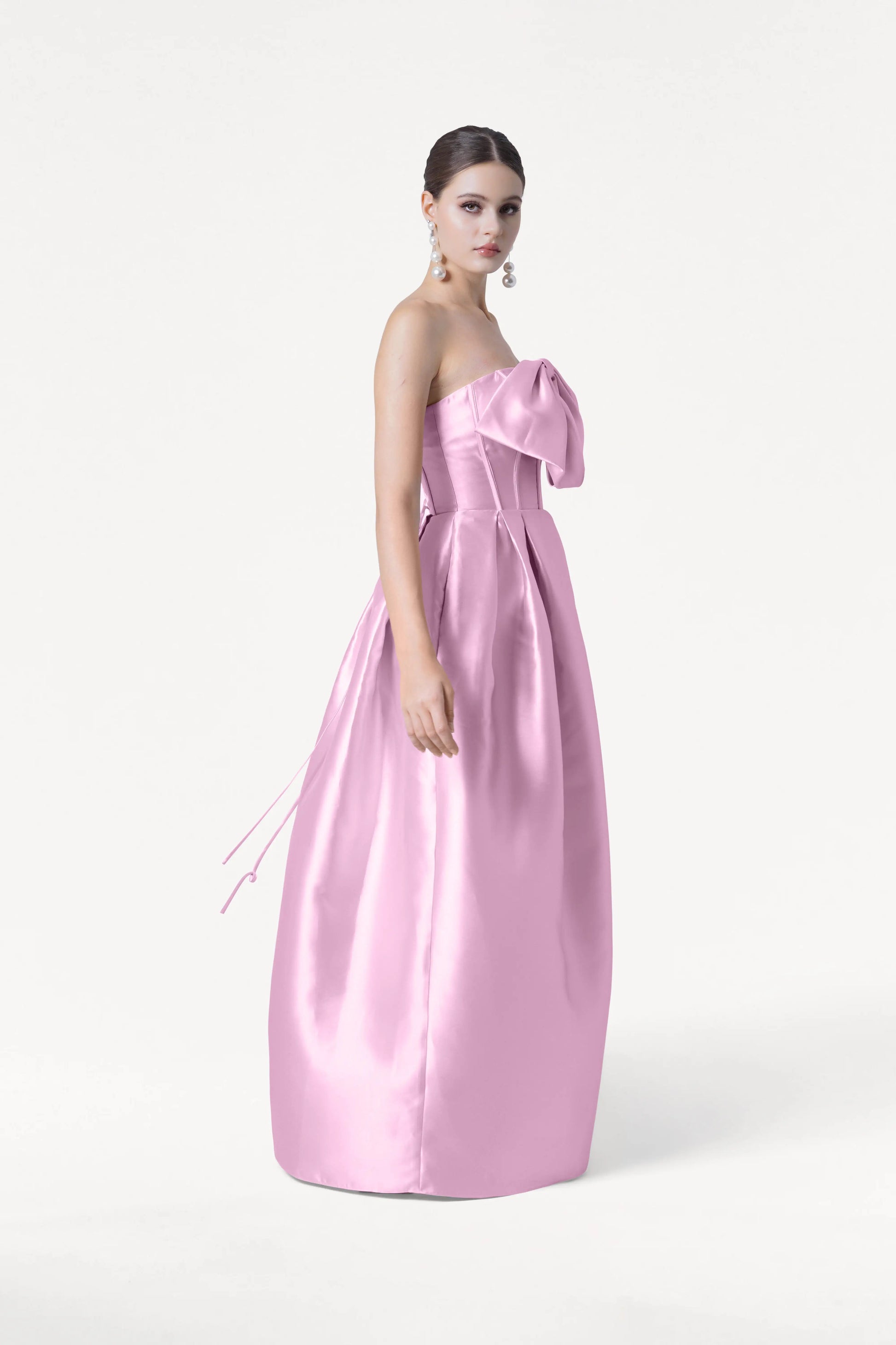 A woman poses against a white background in the TH&TH Ophelia Bridesmaid Dress in bubblegum pink satin twill, featuring a large bow and corset bodice. Her hair is styled in a sleek updo and she wears long, dangling earrings.