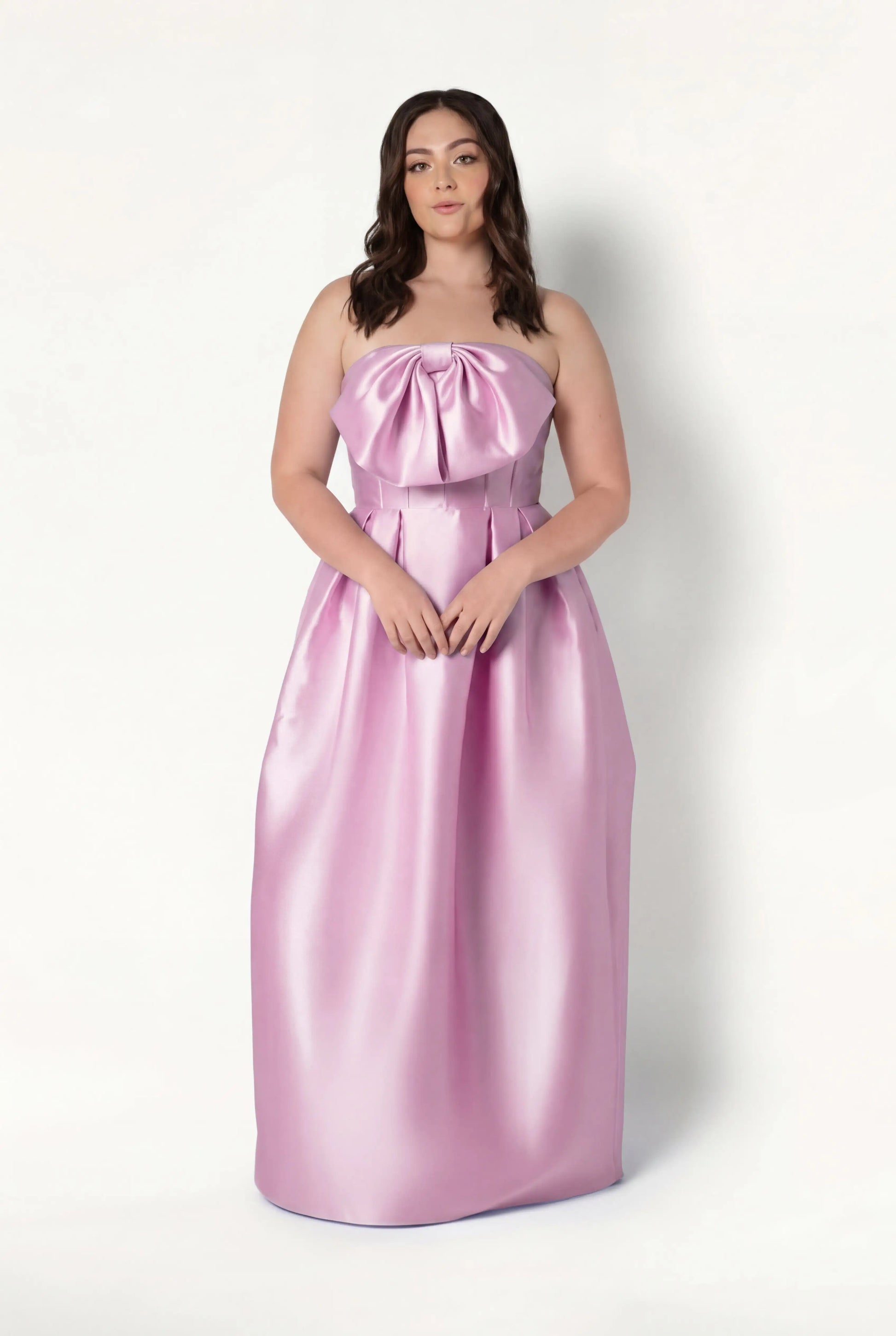 A woman wears the TH&TH Ophelia Bridesmaid Dress in bubblegum pink satin twill—a strapless, floor-length gown with a corset bodice and large bow—standing against a plain white background.