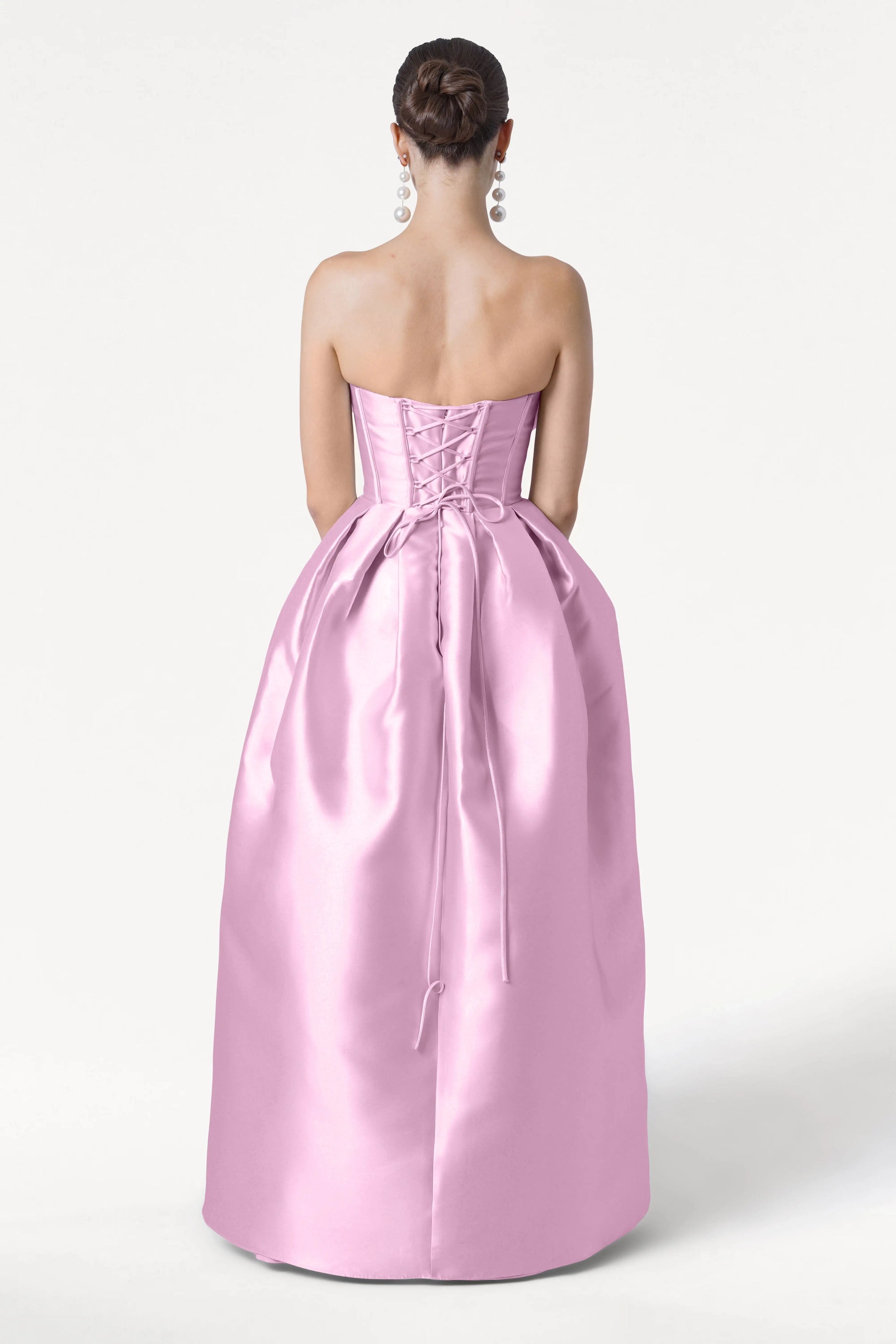 A woman faces away, wearing the TH&TH Ophelia Bridesmaid Dress in Bubblegum Pink Satin Twill, featuring a strapless corset bodice and lace-up back. Her hair is in a neat bun with large dangling earrings, set against a plain white background.
