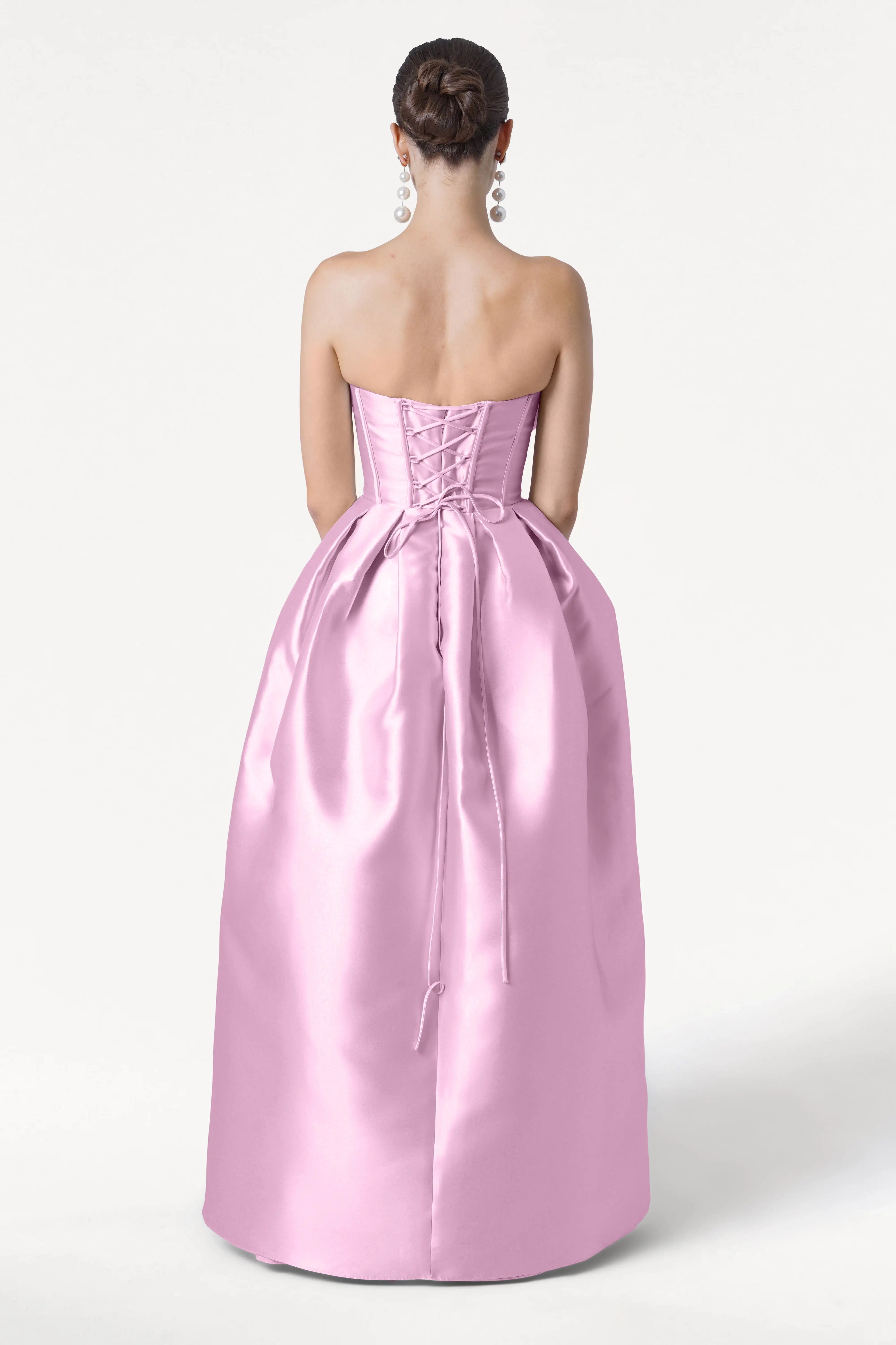 A woman faces away, wearing the TH&TH Ophelia Bridesmaid Dress in Bubblegum Pink Satin Twill, featuring a strapless corset bodice and lace-up back. Her hair is in a neat bun with large dangling earrings, set against a plain white background.