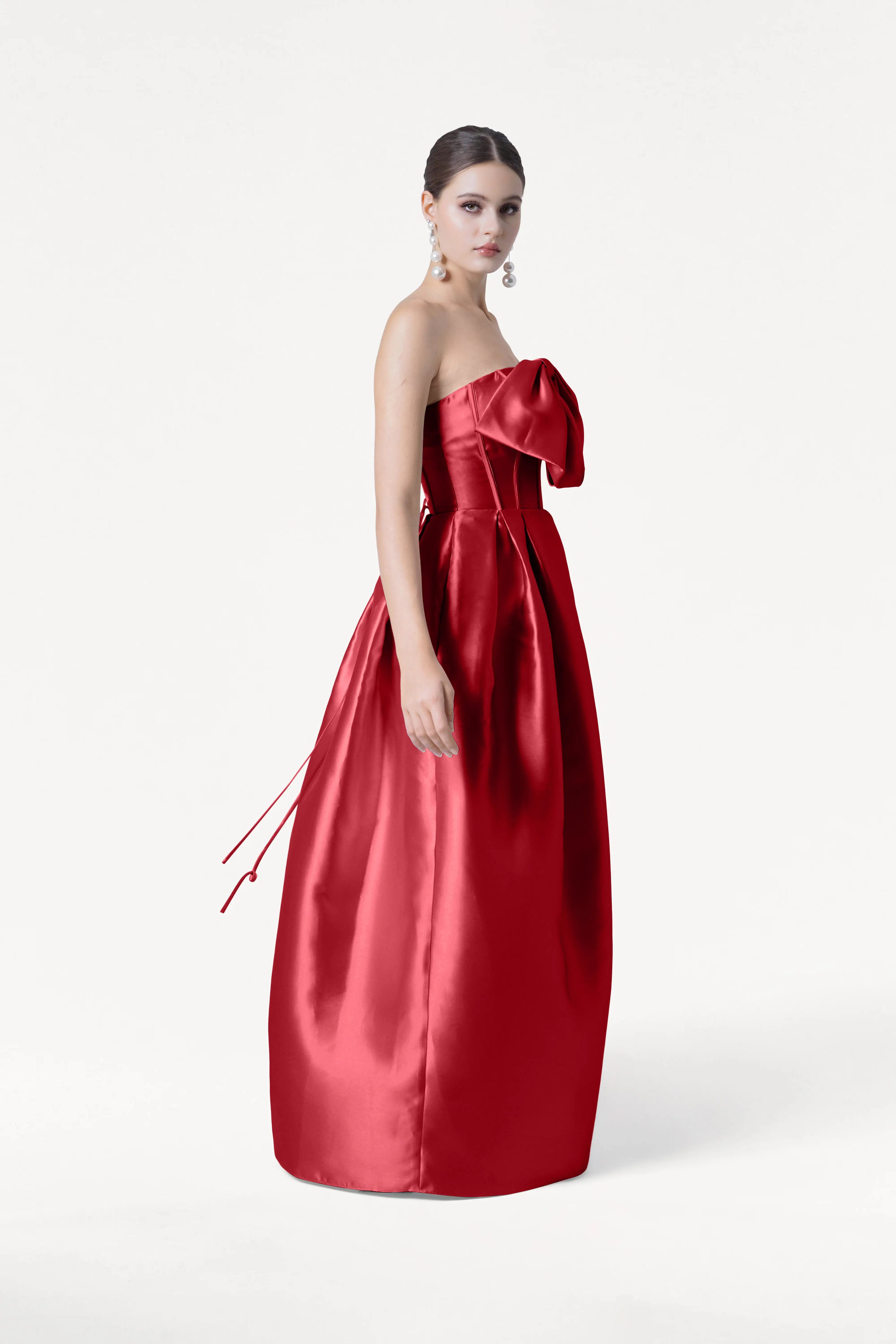 A woman stands in profile wearing the TH&TH Ophelia Bridesmaid Dress in Dark Cherry Satin Twill, featuring a strapless corset bodice and dramatic bow. Her hair is styled in a sleek updo with long, dangling earrings against a plain white background.