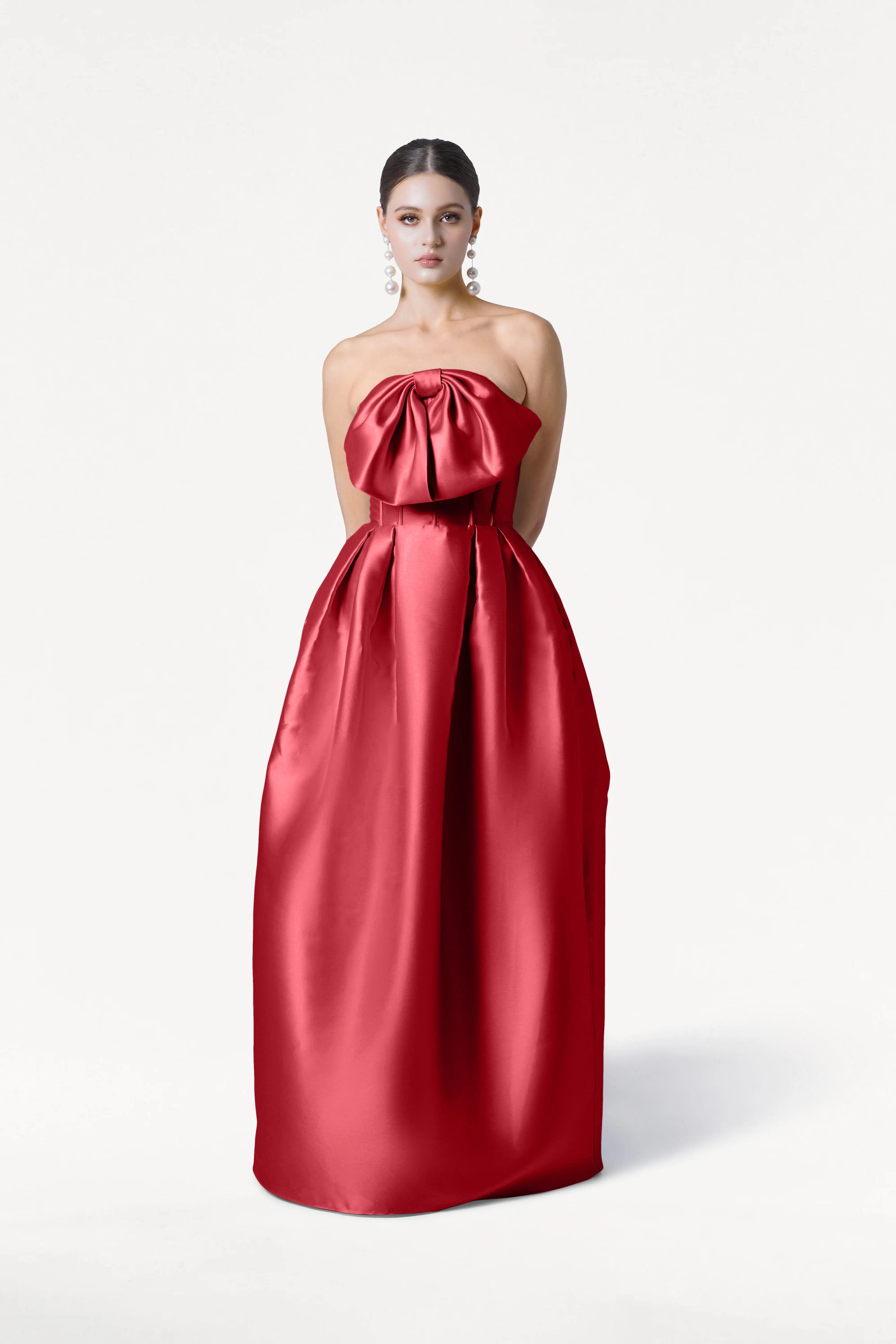 A woman stands against a white background wearing the Ophelia Bridesmaid Dress in Dark Cherry Satin Twill by TH&TH, featuring a strapless, floor-length silhouette with a large bow at the bust. She has her hands behind her back and wears statement drop earrings.
