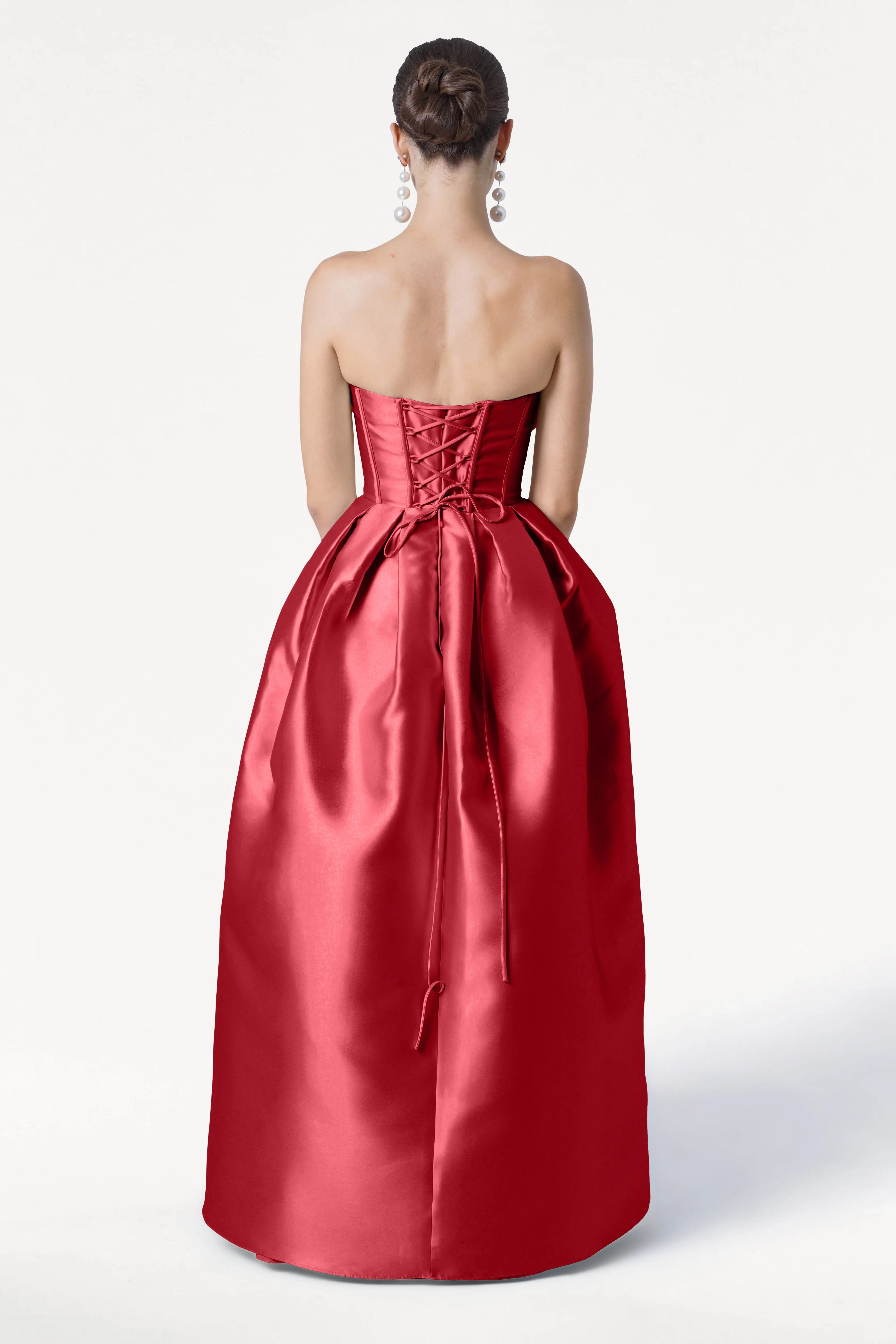 A woman with an updo and long earrings stands against a plain white background, wearing the TH&TH Ophelia Bridesmaid Dress in Dark Cherry Satin Twill. The strapless gown features a corset bodice and a full skirt.