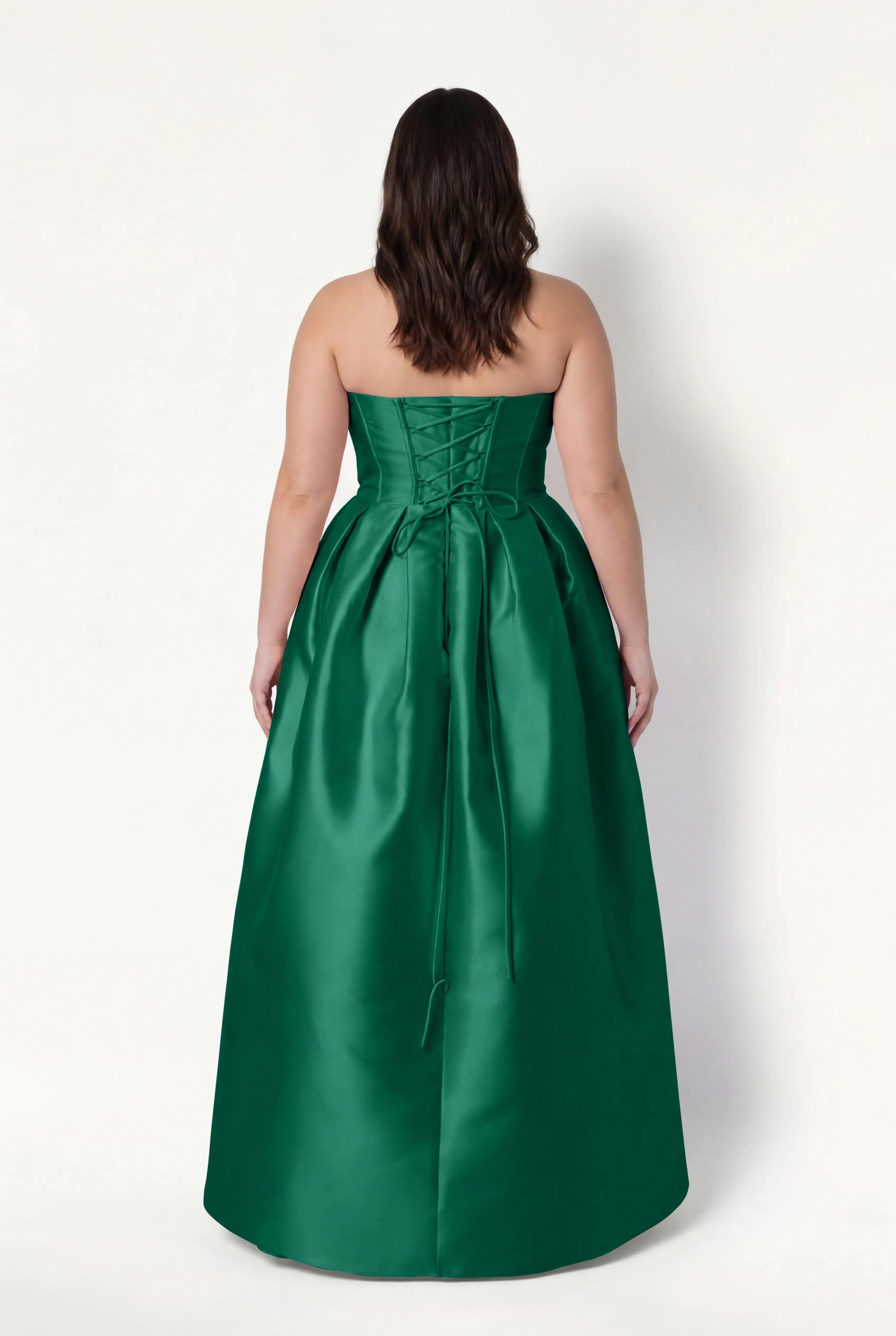 A woman with wavy brown hair is seen from the back wearing the TH&TH Ophelia Bridesmaid Dress in Forest Green Satin Twill, a strapless floor-length design with a corset bodice, waist bow, and smooth satin twill finish.