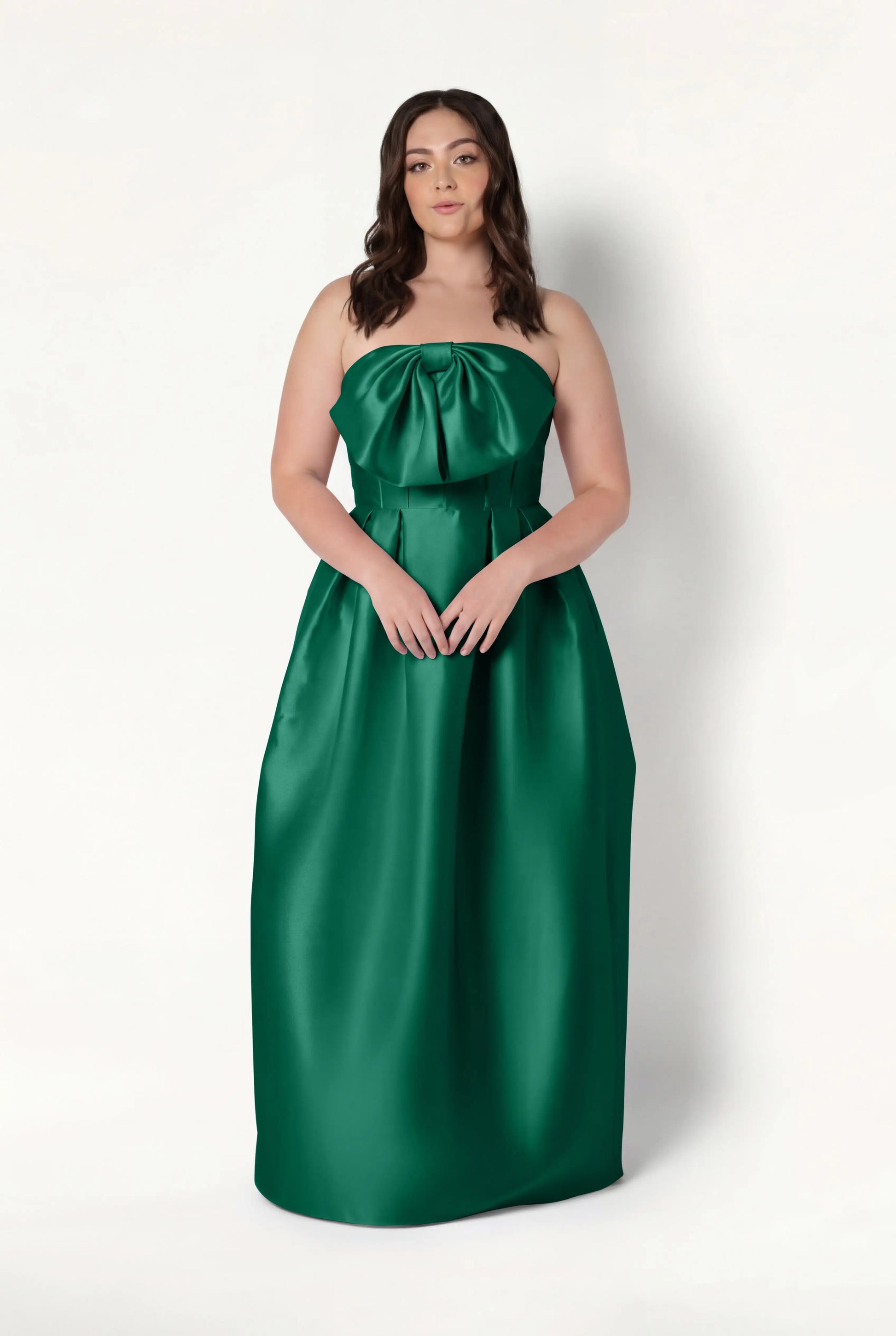 A woman with long brown hair wears the TH&TH Ophelia Bridesmaid Dress in Forest Green Satin Twill, featuring a strapless corset bodice and large bow detail, standing against a plain white background.