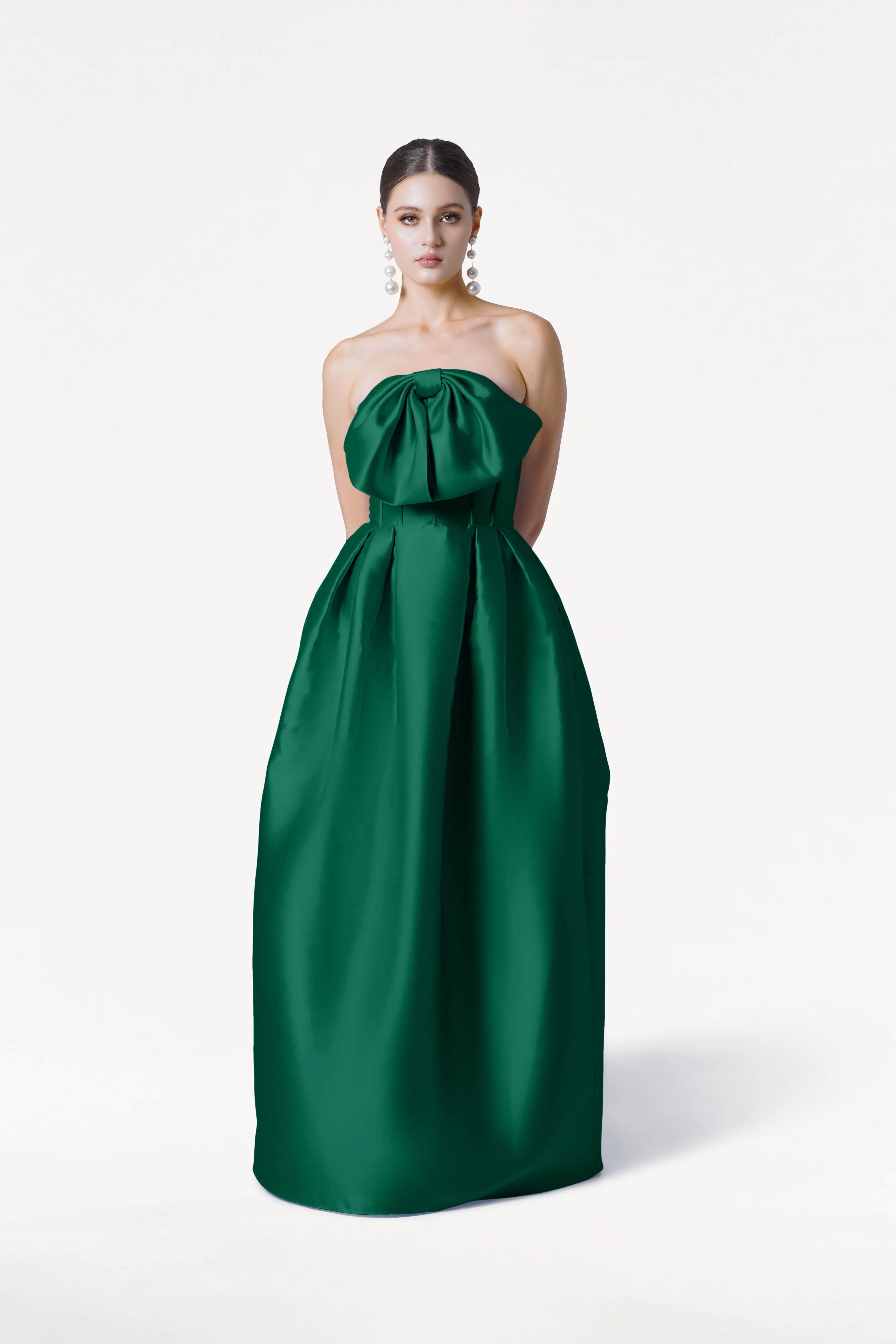 A woman with her hair pulled back and statement earrings wears the TH&TH Ophelia Bridesmaid Dress in Forest Green Satin Twill, a strapless, floor-length gown with a large bow detail on the corset bodice, against a white background.