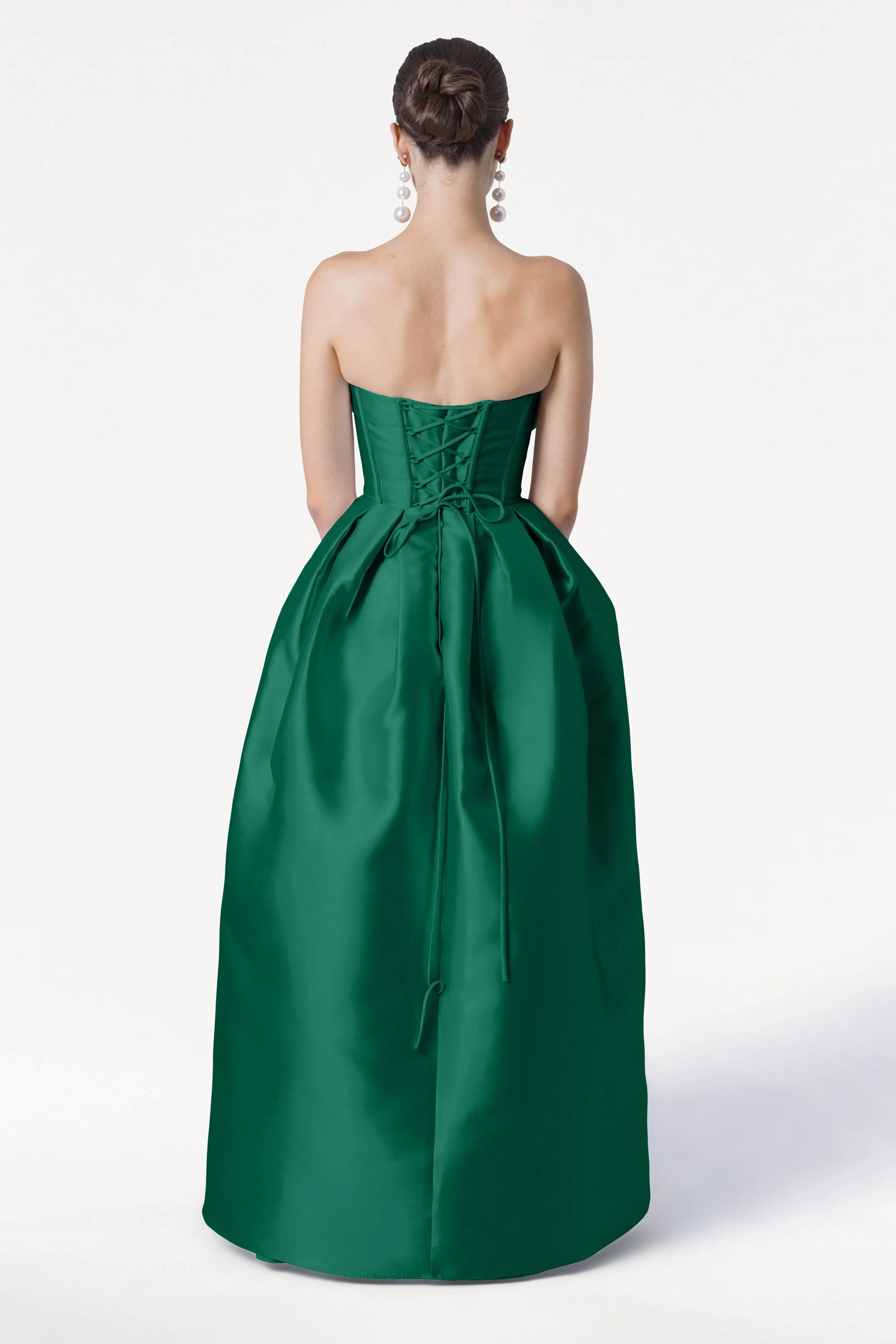 A woman in the TH&TH Ophelia Bridesmaid Dress in Forest Green Satin Twill, featuring a strapless corset bodice and lace-up back, stands facing away. Her hair is in a neat bun, with large dangling earrings against a plain white background.