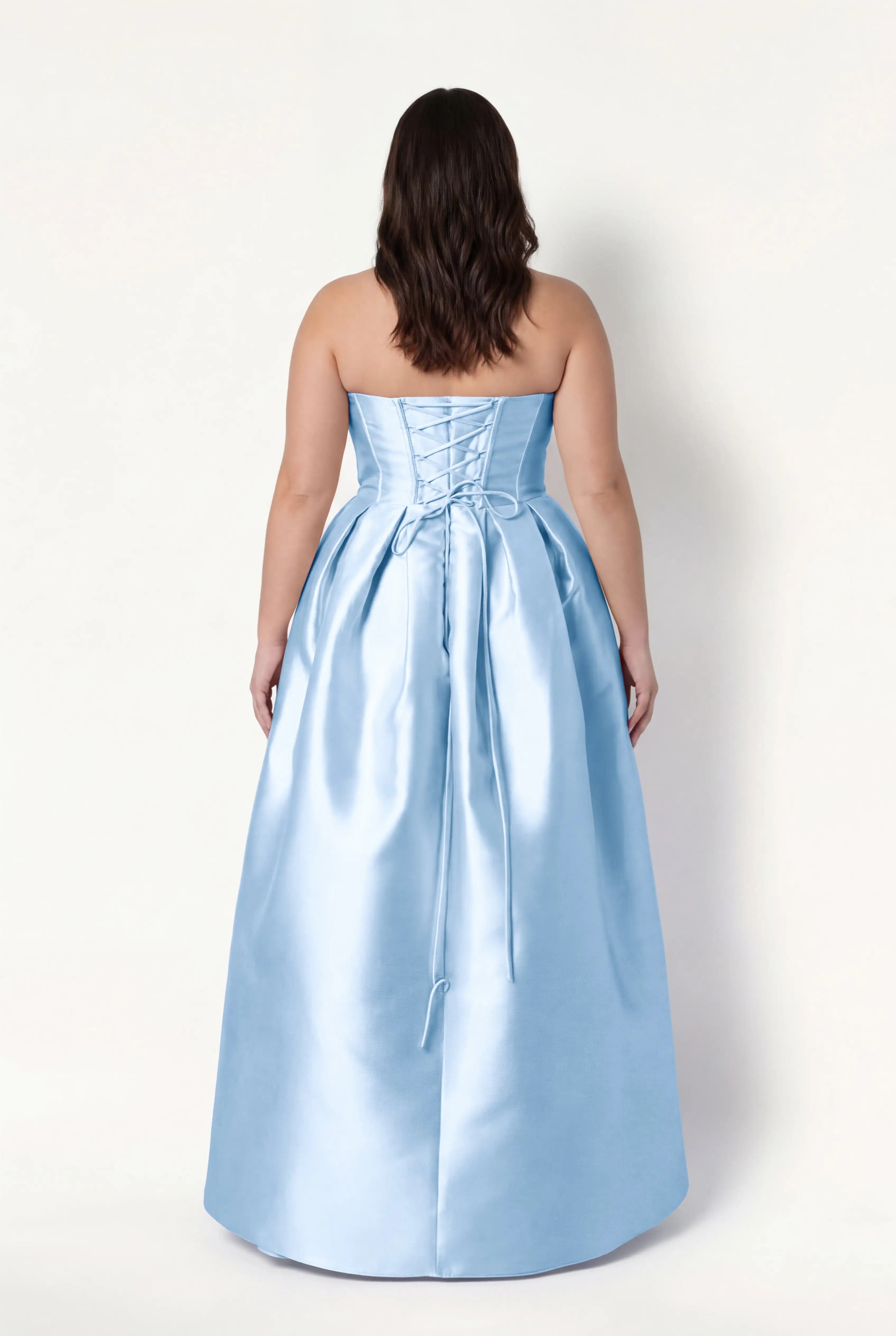 A woman with wavy, shoulder-length brown hair models the TH&TH Ophelia Bridesmaid Dress in Frosted Blue Satin Twill, featuring a strapless lace-up corset bodice, standing with her back to the camera against a plain white background.