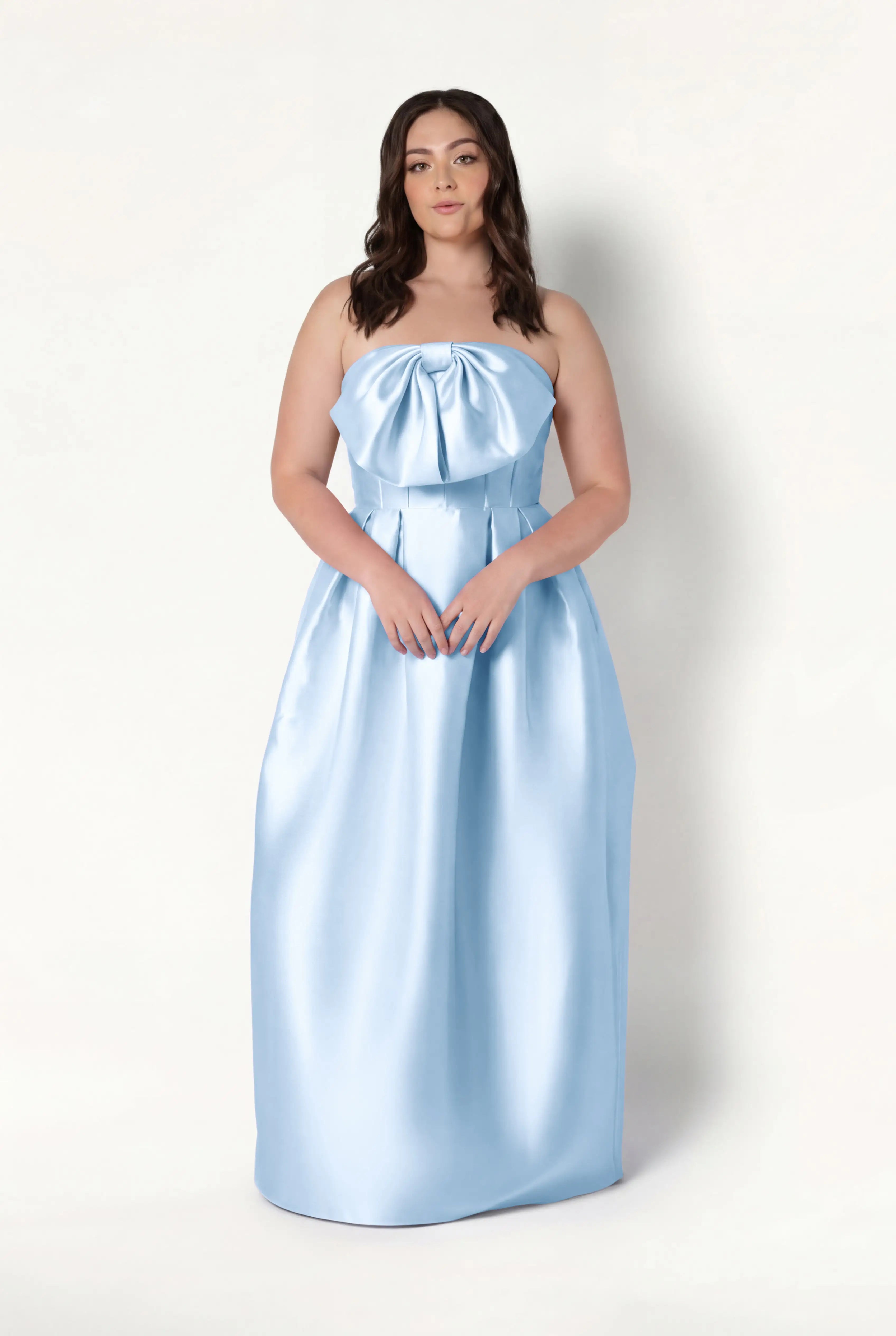 A woman with long brown hair models the TH&TH Ophelia Bridesmaid Dress in Frosted Blue Satin Twill, a strapless floor-length gown with a large bow and corset bodice, standing against a plain white background.