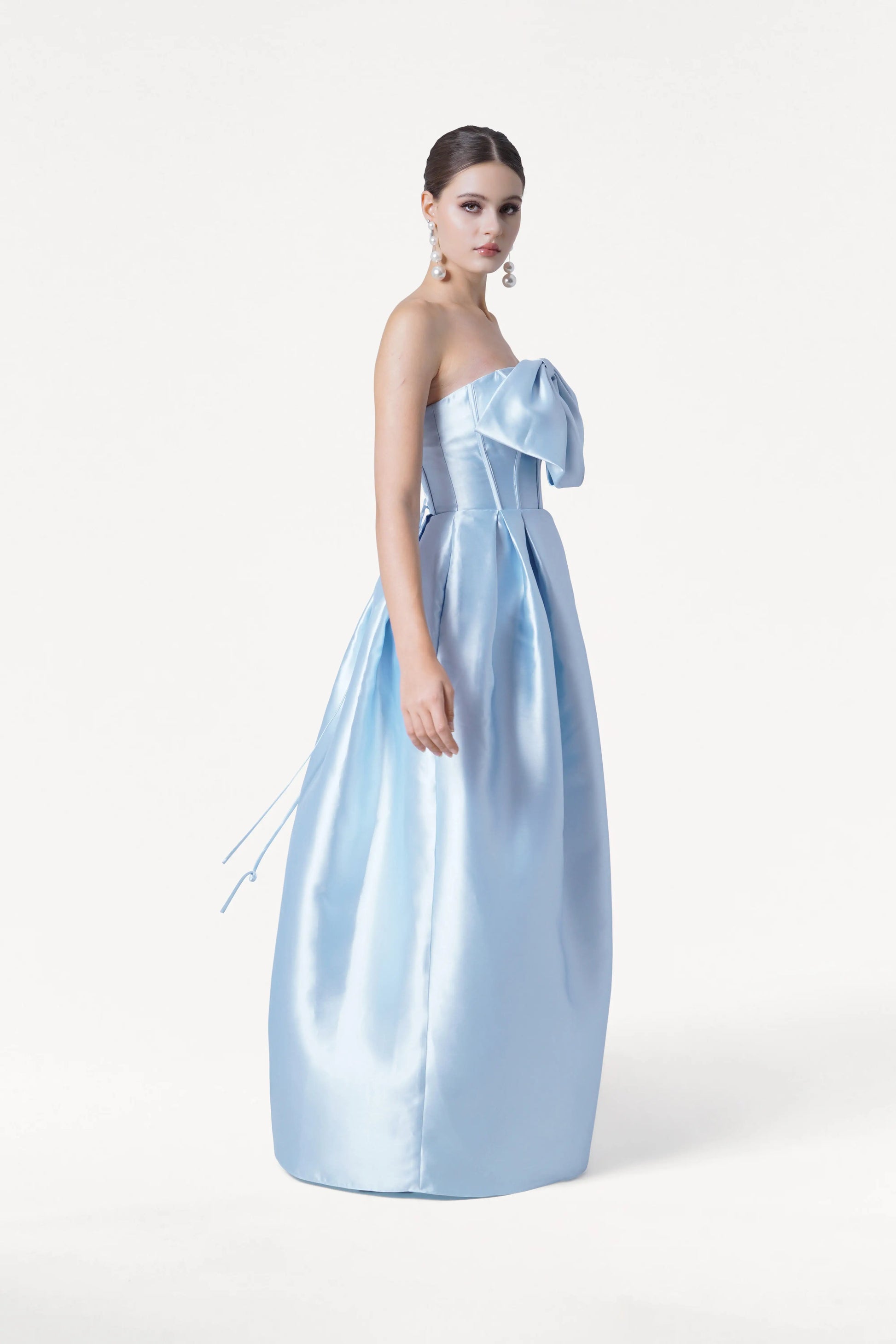 A woman models the TH&TH Ophelia Bridesmaid Dress in Frosted Blue Satin Twill, featuring a strapless corset bodice and large bow detail. She wears dangling earrings, her hair in a sleek updo, against a plain white background.