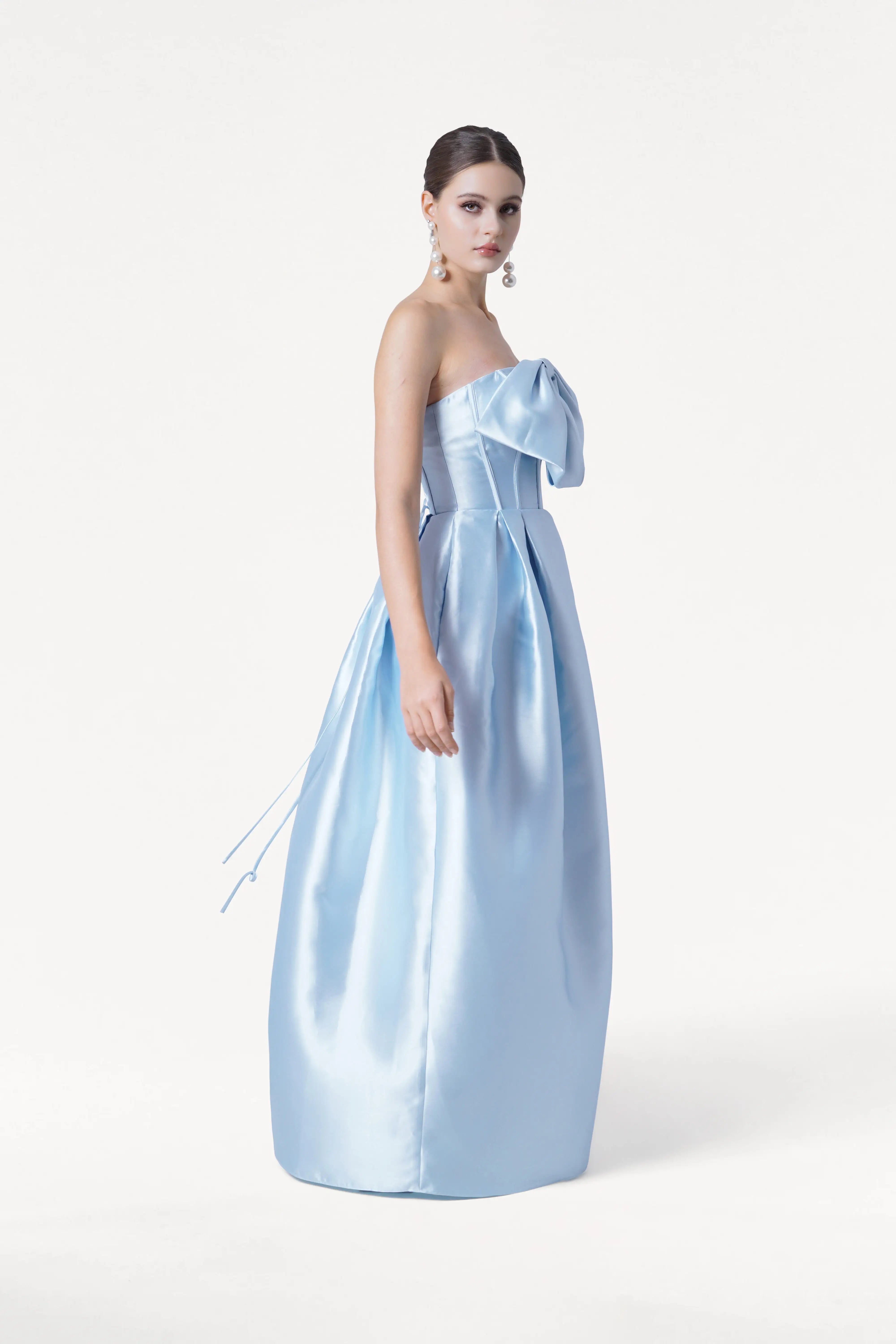 A woman models the TH&TH Ophelia Bridesmaid Dress in Frosted Blue Satin Twill, featuring a strapless corset bodice and large bow detail. She wears dangling earrings, her hair in a sleek updo, against a plain white background.