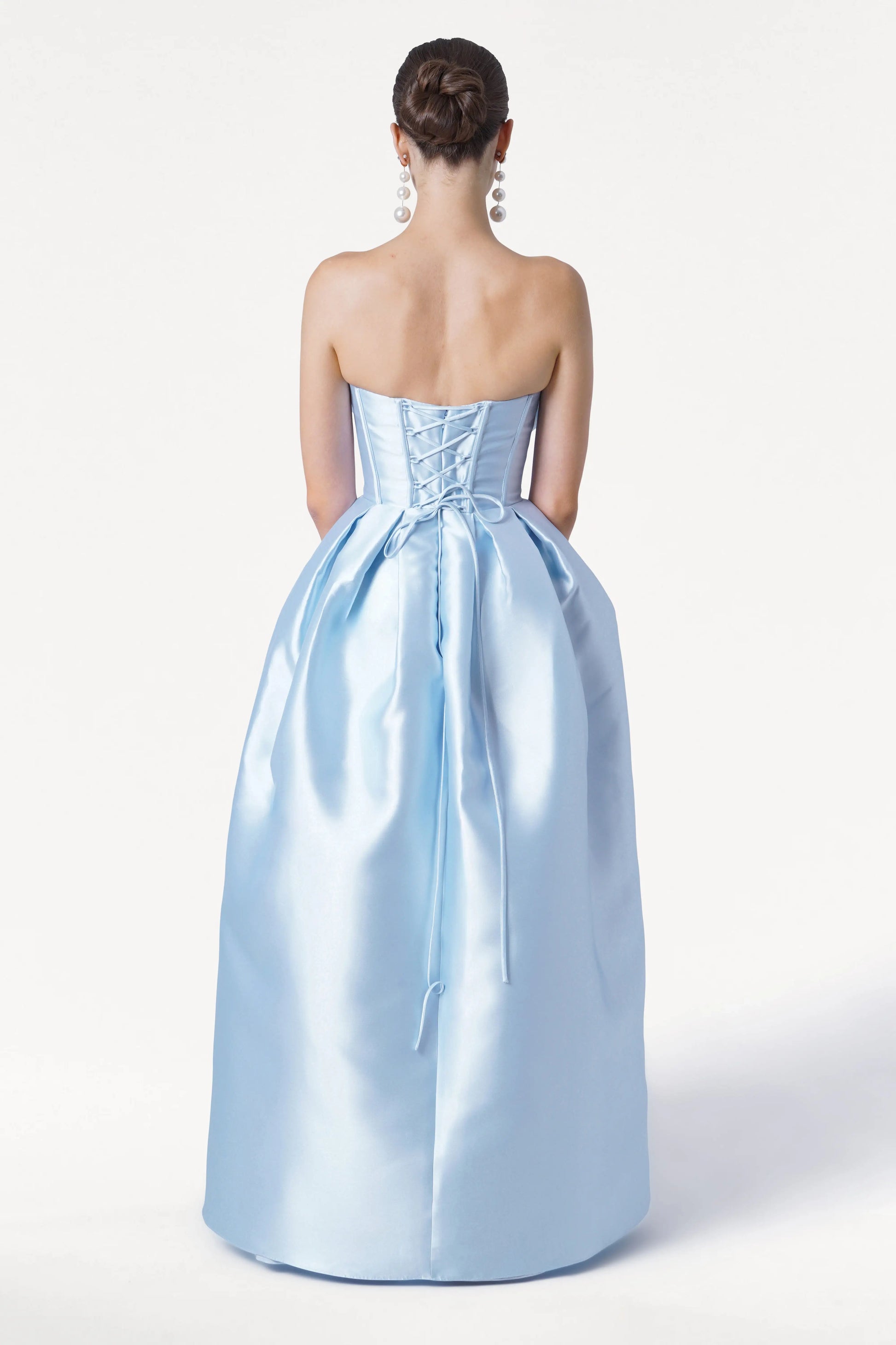 A woman with her hair in a bun is seen from behind wearing the TH&TH Ophelia Bridesmaid Dress in Frosted Blue Satin Twill, featuring a strapless lace-up corset bodice and full skirt. She accessorizes with long, dangling earrings against a plain white background.