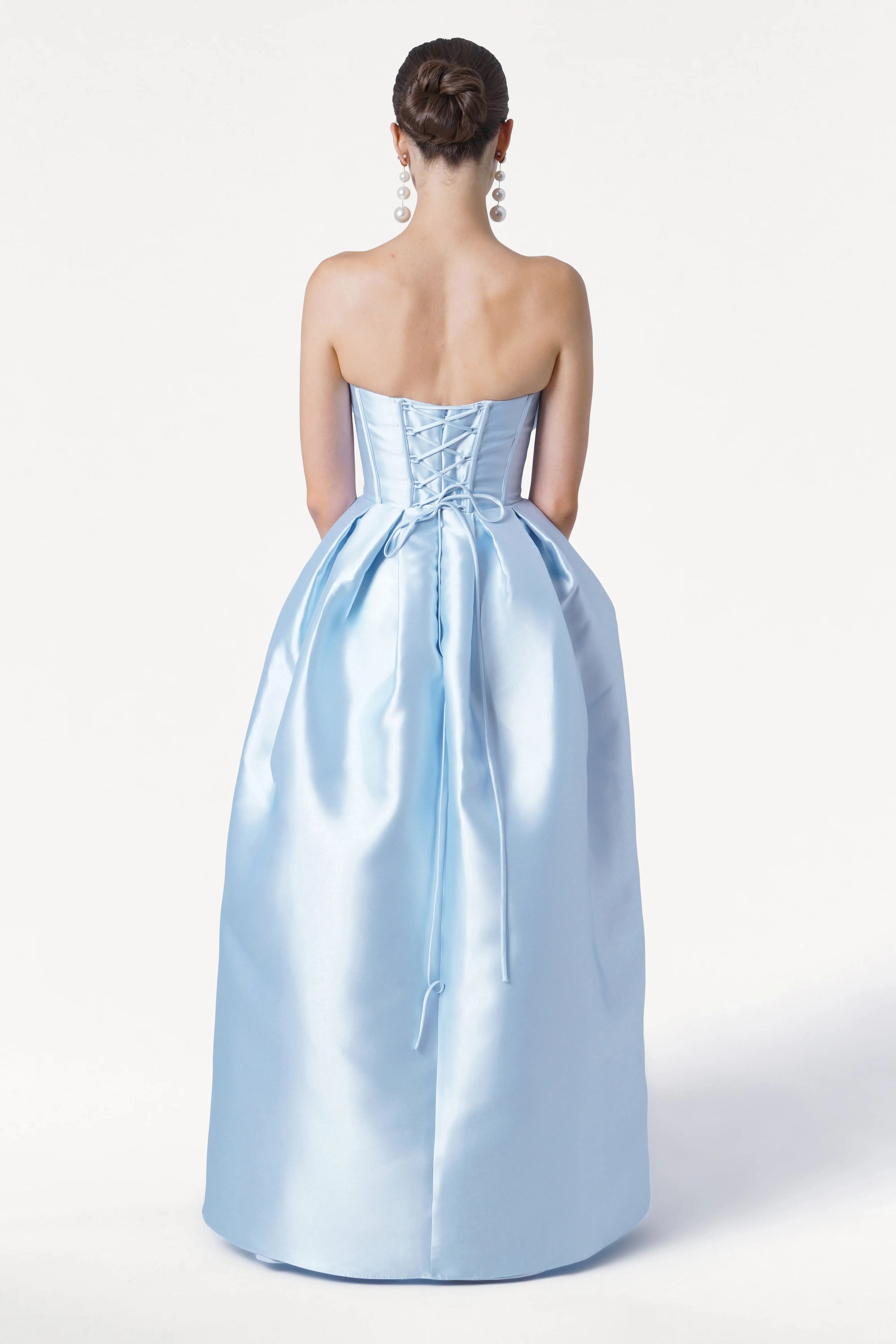 A woman with her hair in a bun is seen from behind wearing the TH&TH Ophelia Bridesmaid Dress in Frosted Blue Satin Twill, featuring a strapless lace-up corset bodice and full skirt. She accessorizes with long, dangling earrings against a plain white background.