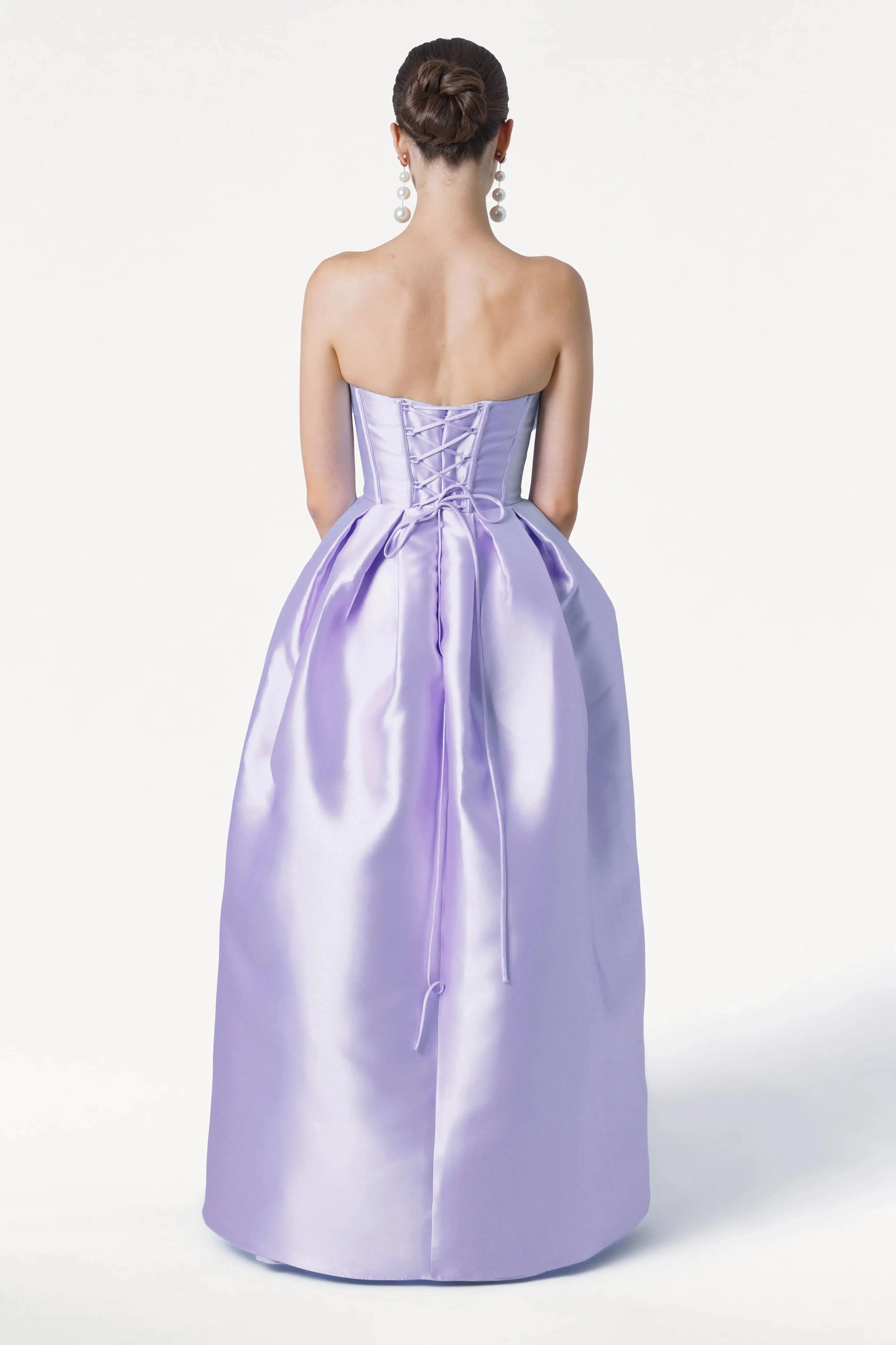 A woman with a neat bun and dangling earrings faces away, modeling the TH&TH Ophelia Bridesmaid Dress in Lilac Mist Satin Twill, featuring a strapless lace-up corset bodice and floor-length skirt against a plain white background.