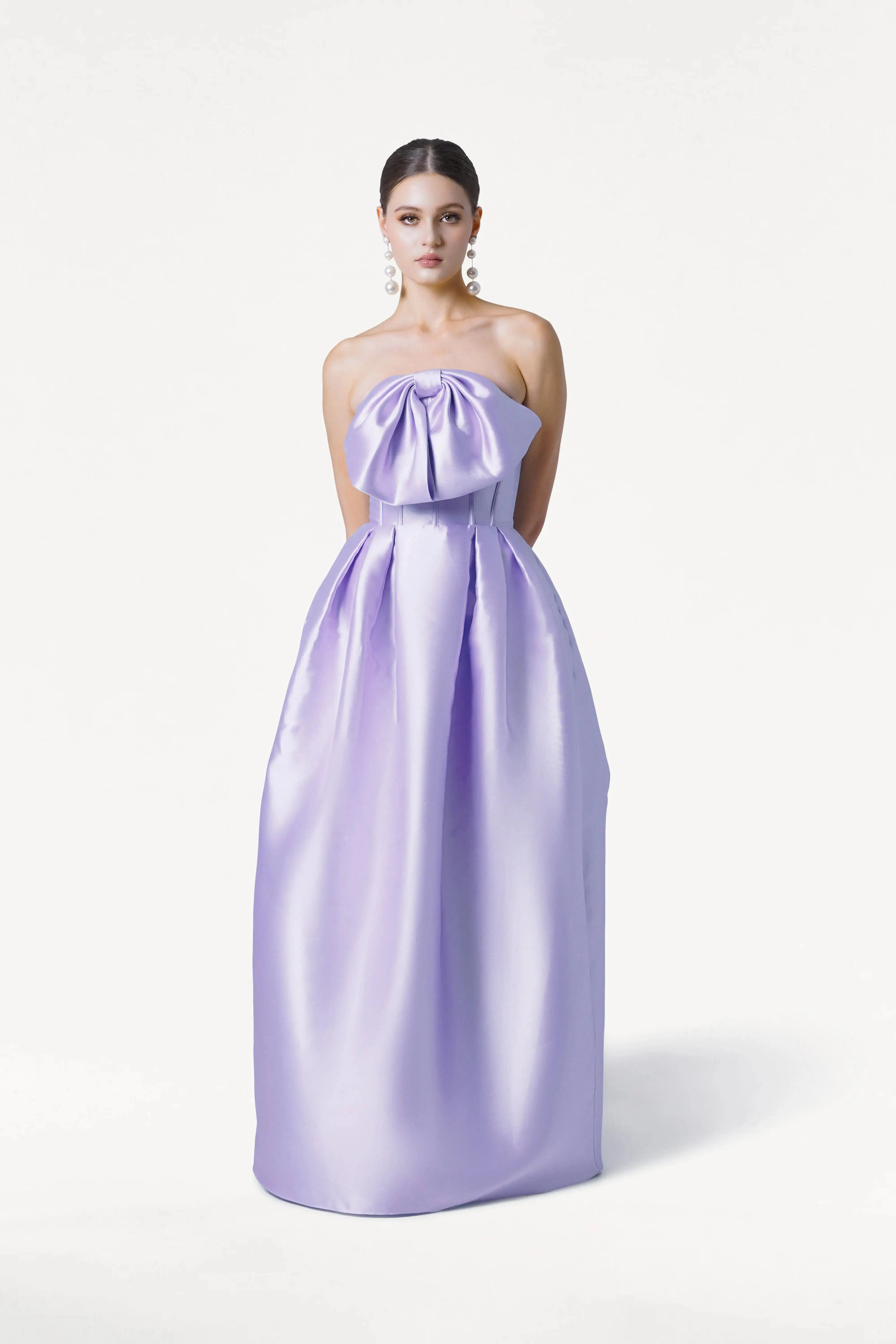 A woman poses against a white backdrop wearing the TH&TH Ophelia Bridesmaid Dress in Lilac Mist Satin Twill, a strapless, floor-length gown with a corset bodice and large bow. Her hair is styled back, and she accessorizes with dangling pearl earrings.