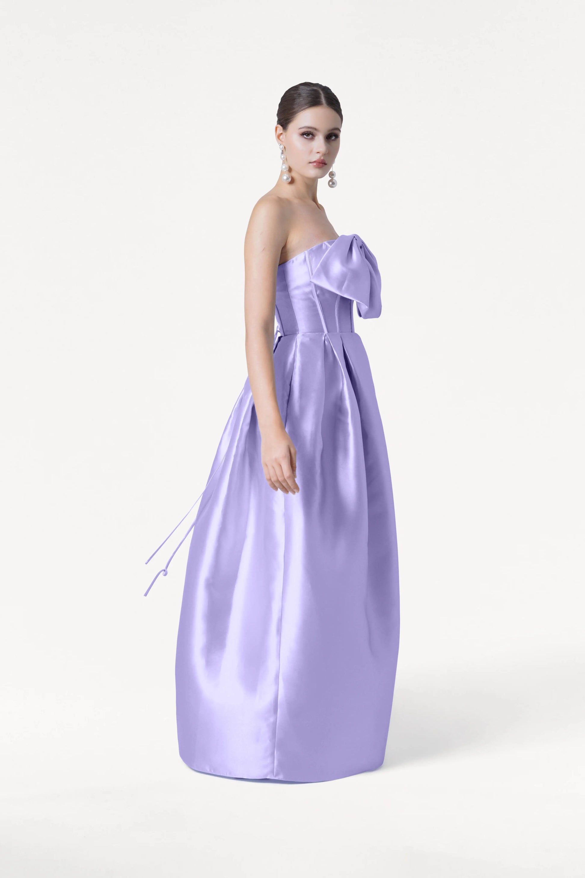 A woman models the TH&TH Ophelia Bridesmaid Dress in Lilac Mist Satin Twill—a strapless, floor-length gown with a corset bodice and large bow—against a white background, styled with statement earrings and an elegant updo.