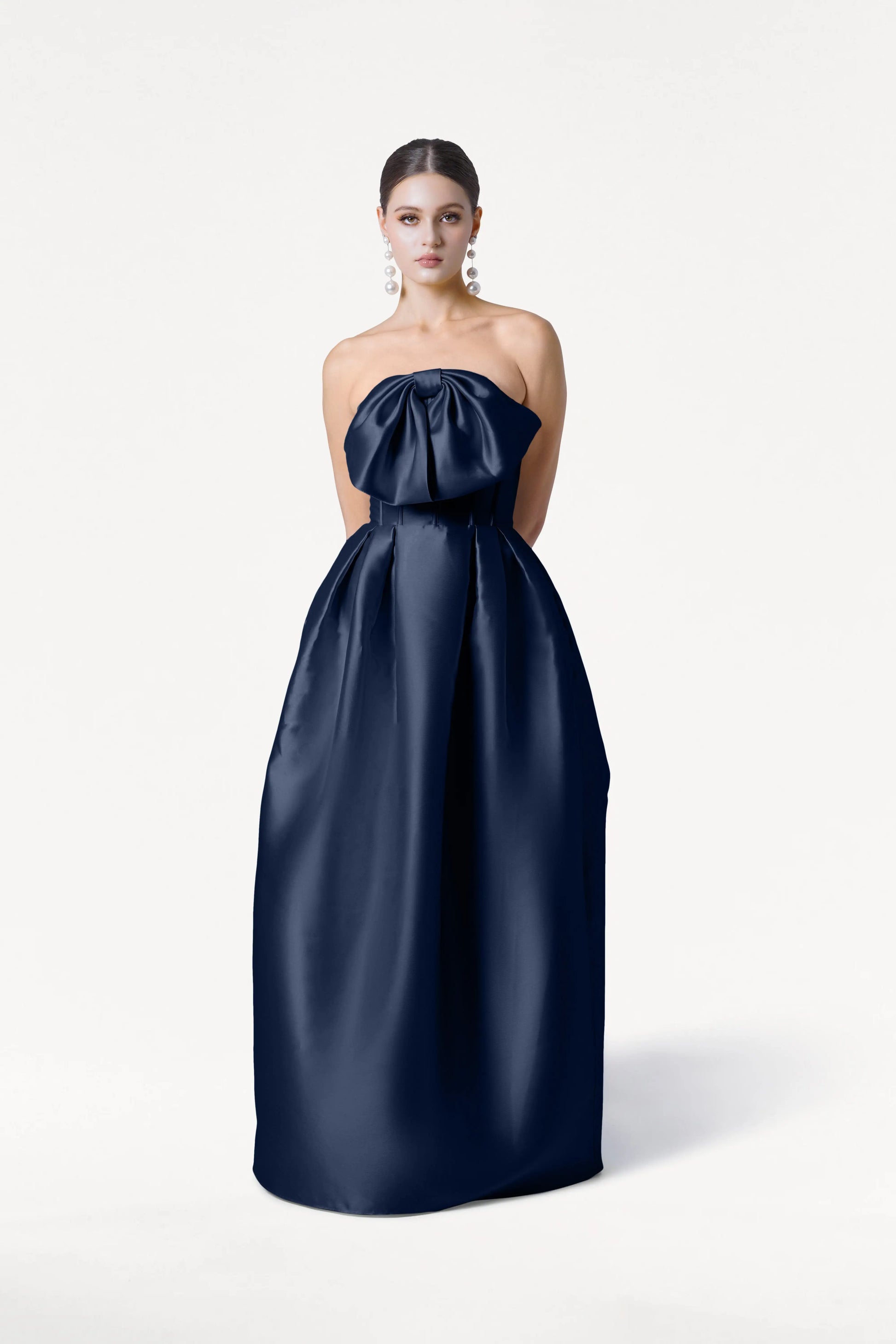 A woman models the TH&TH Ophelia Bridesmaid Dress in Navy Satin Twill, a strapless, floor-length gown featuring a large bow at the chest. She accessorizes with long earrings and her hair is pulled back, standing against a plain white background.