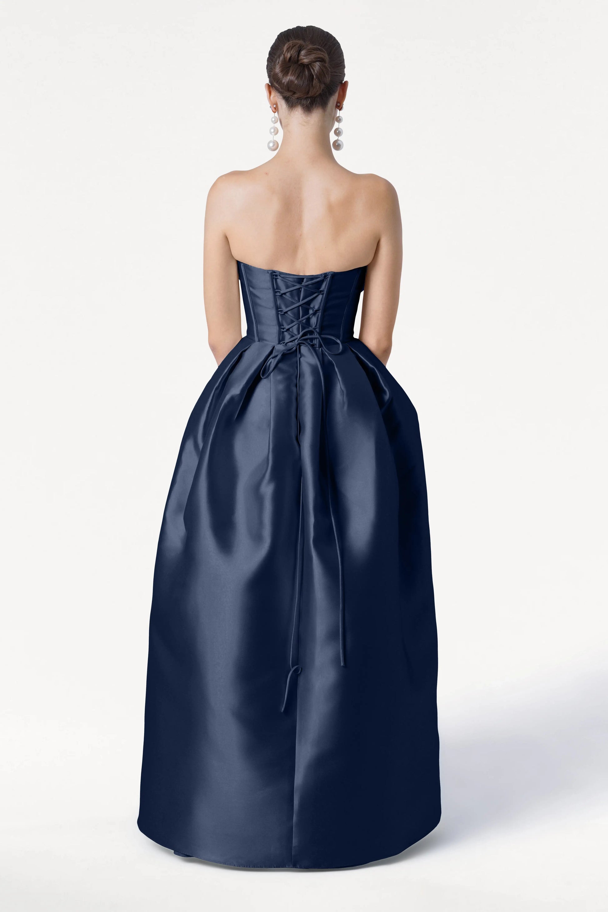 A woman with an elegant updo wears the TH&TH Ophelia Bridesmaid Dress in Navy Satin Twill, featuring a strapless corset bodice, lace-up back, and waist bow. She accessorizes with large dangling earrings against a plain white background.