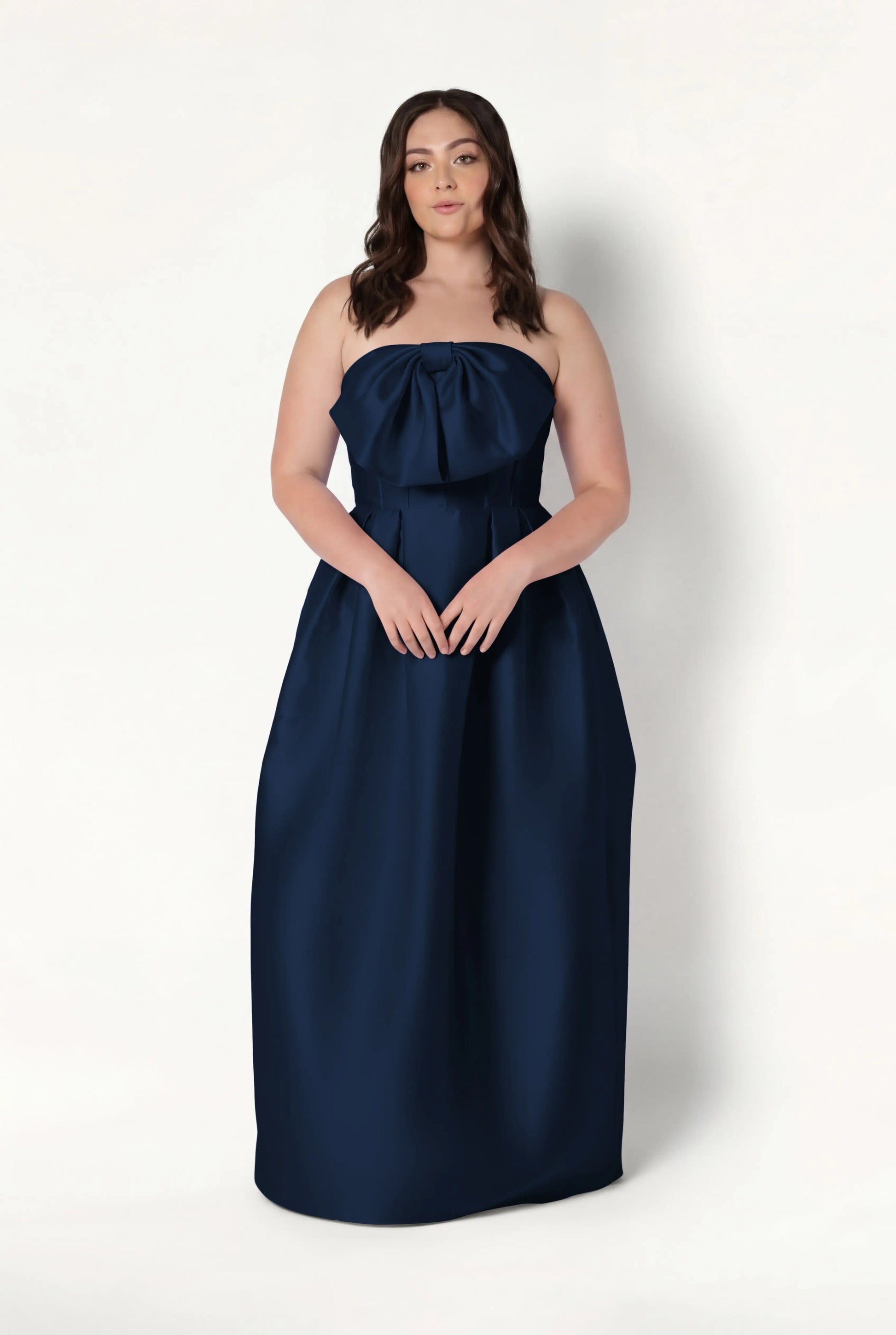 A woman with wavy brown hair models the TH&TH Ophelia Bridesmaid Dress in navy satin twill, a strapless, floor-length gown with a large bow and corset bodice, against a plain white background.