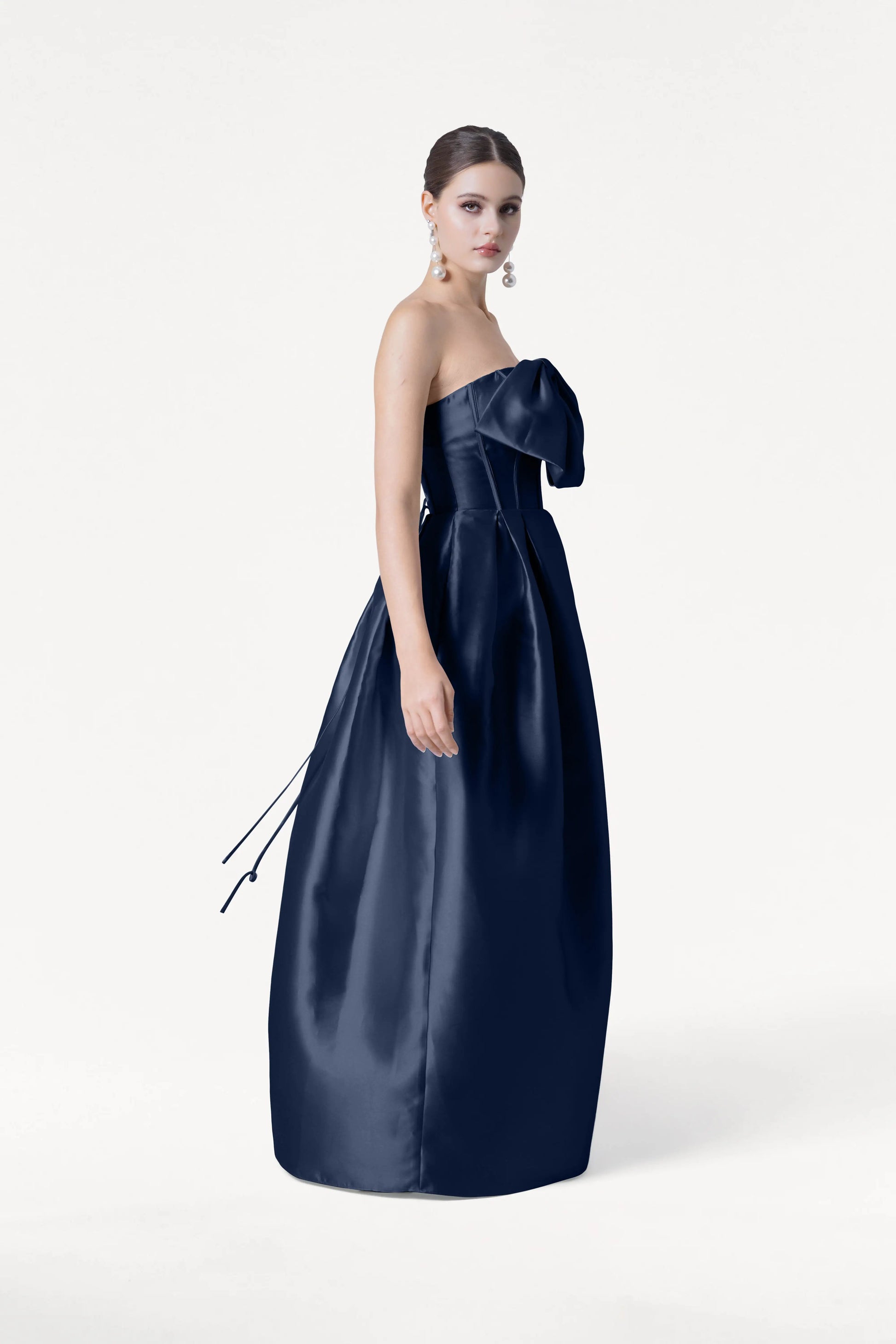 A woman stands against a white background wearing the TH&TH Ophelia Bridesmaid Dress in Navy Satin Twill, which features a strapless corset bodice with a large bow. Her hair is styled in an updo and she wears long earrings.