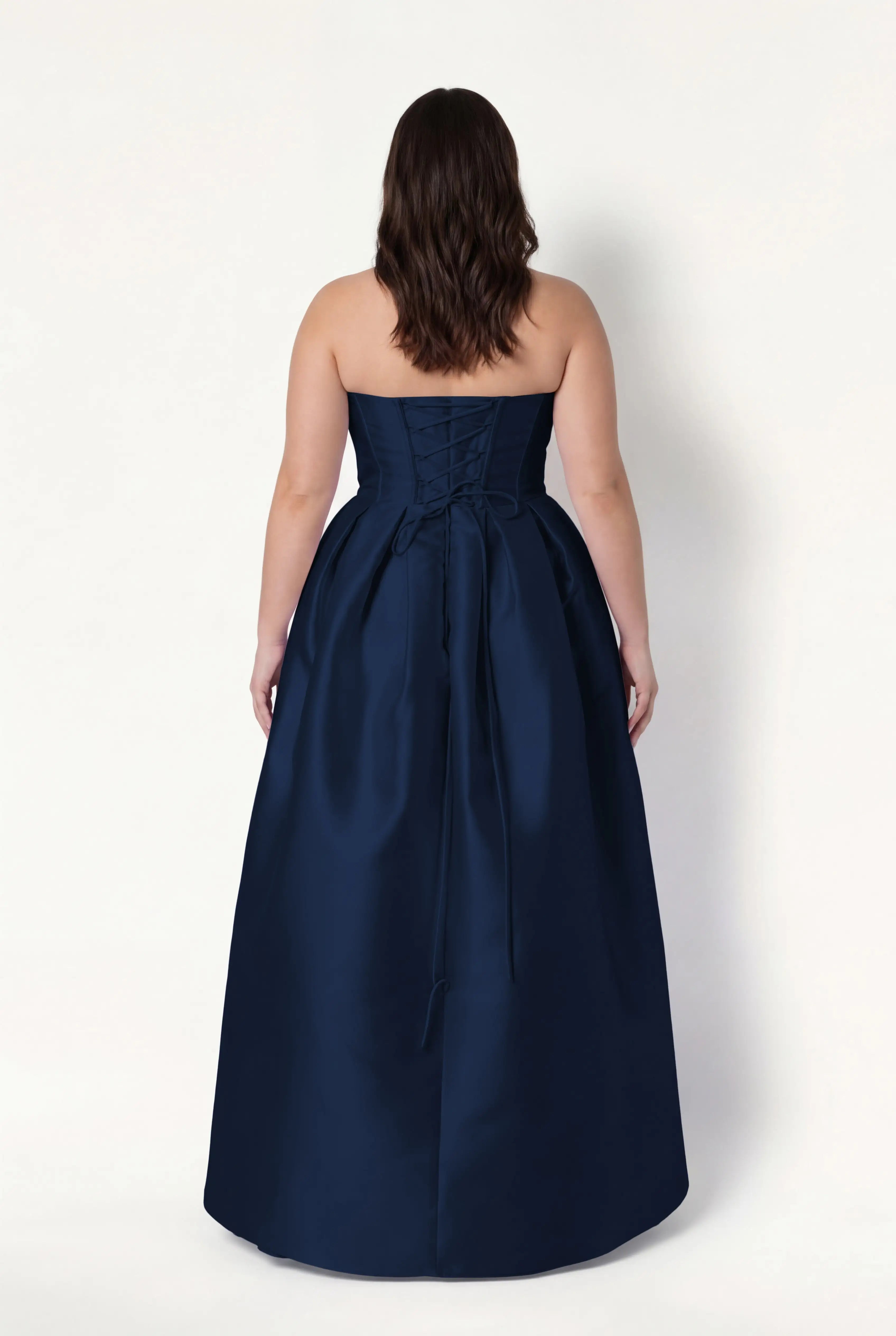 A woman with wavy brown hair stands facing away, wearing the TH&TH Ophelia Bridesmaid Dress in Navy Satin Twill, which features a strapless corset bodice and a long flowing skirt, set against a plain light background.