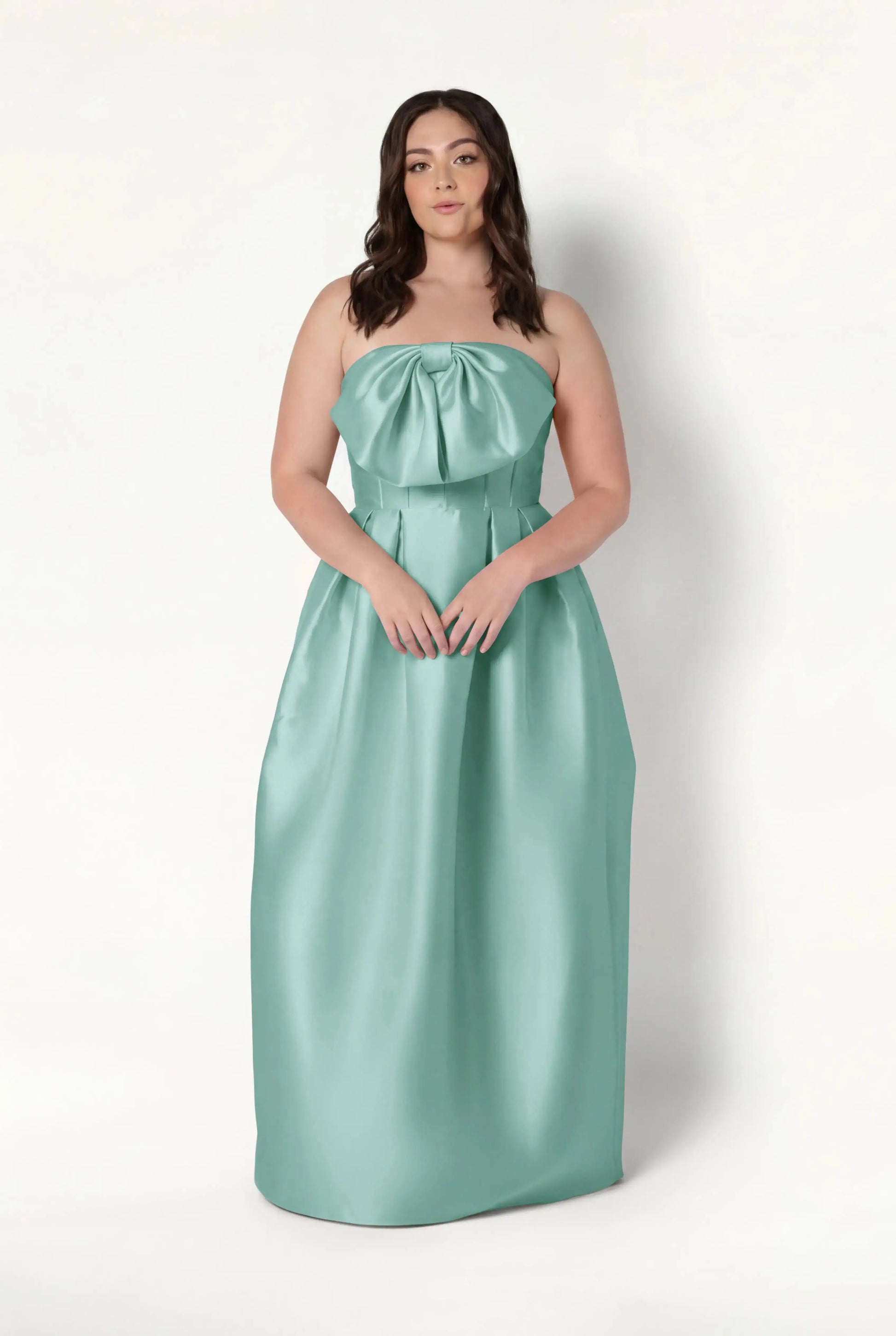 A woman with long, wavy brown hair and a neutral expression wears the TH&TH Ophelia Bridesmaid Dress in Seafoam Satin Twill—a strapless, floor-length gown featuring a corset bodice and large bow—against a plain white background.