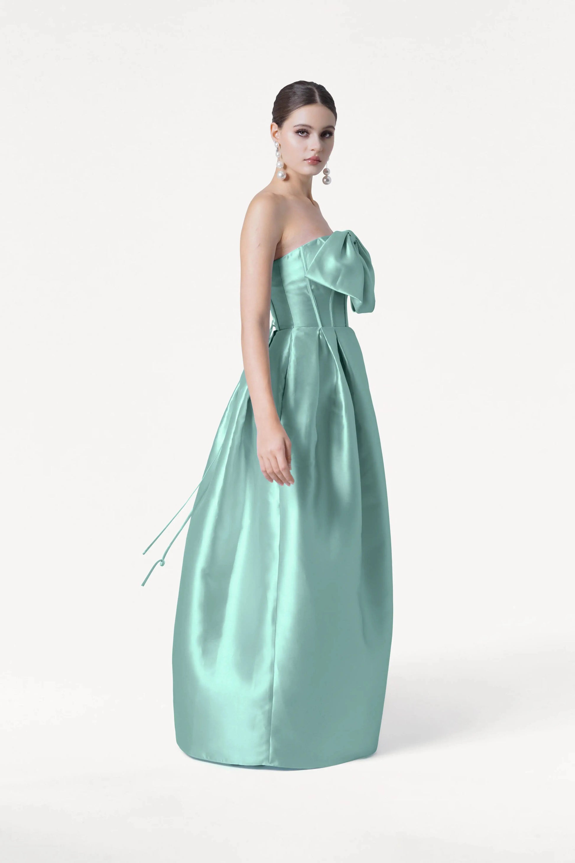 A woman stands sideways against a white background in the TH&TH Ophelia Bridesmaid Dress in Seafoam Satin Twill—a strapless, floor-length gown with a large chest bow. Her hair is tied back and she wears long, dangling earrings.