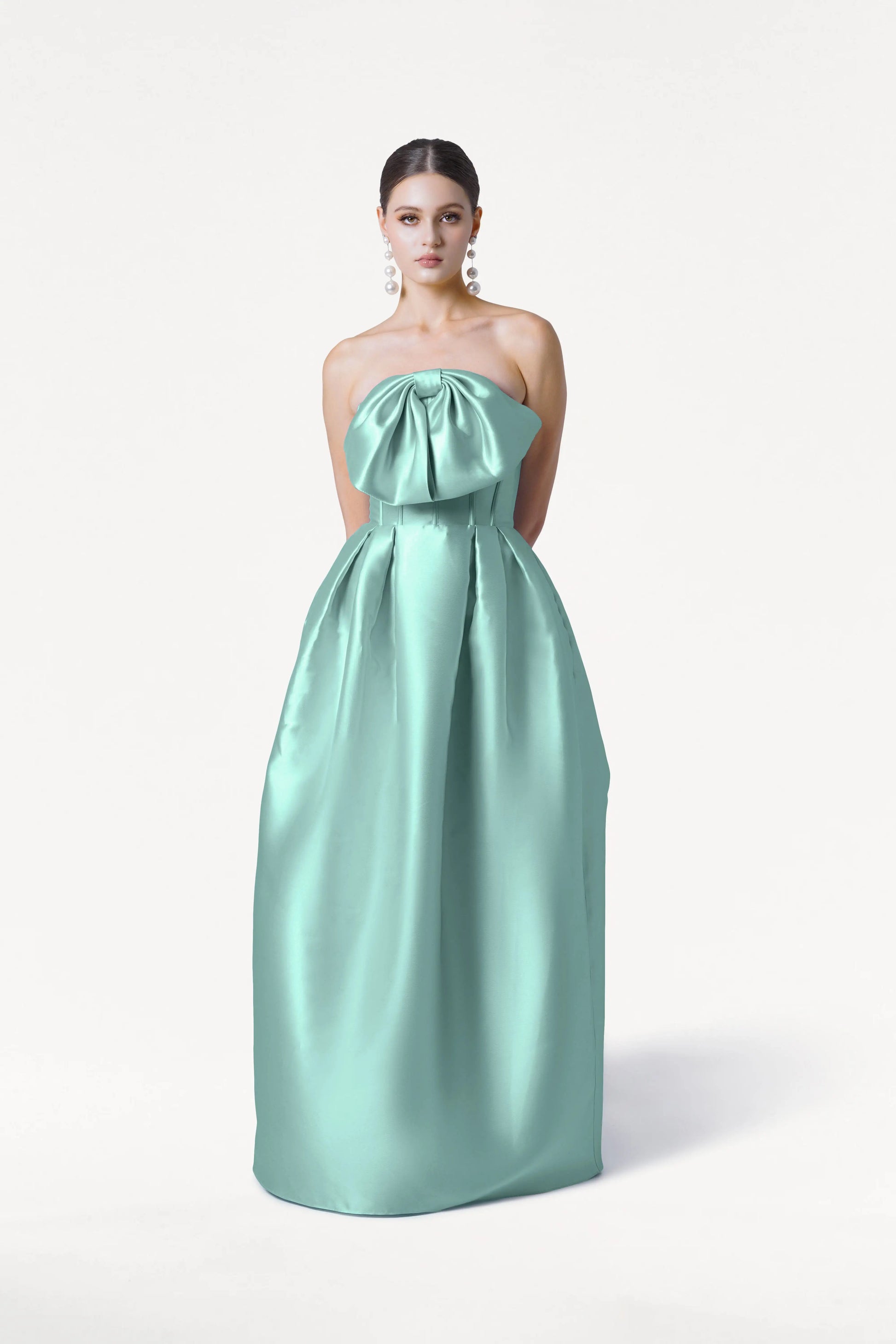 A woman stands against a white background wearing the TH&TH Ophelia Bridesmaid Dress in Seafoam Satin Twill, a strapless floor-length gown with a large bow on the corset bodice. Her arms are behind her back and she wears statement earrings.